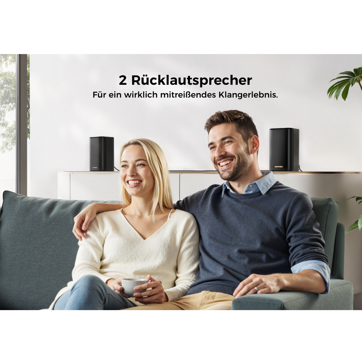Ein Paar lächelt auf einem Sofa. Zwei schwarze Lautsprecher stehen auf einem Regal. Der Hintergrund zeigt ein Fenster und eine Pflanze.
