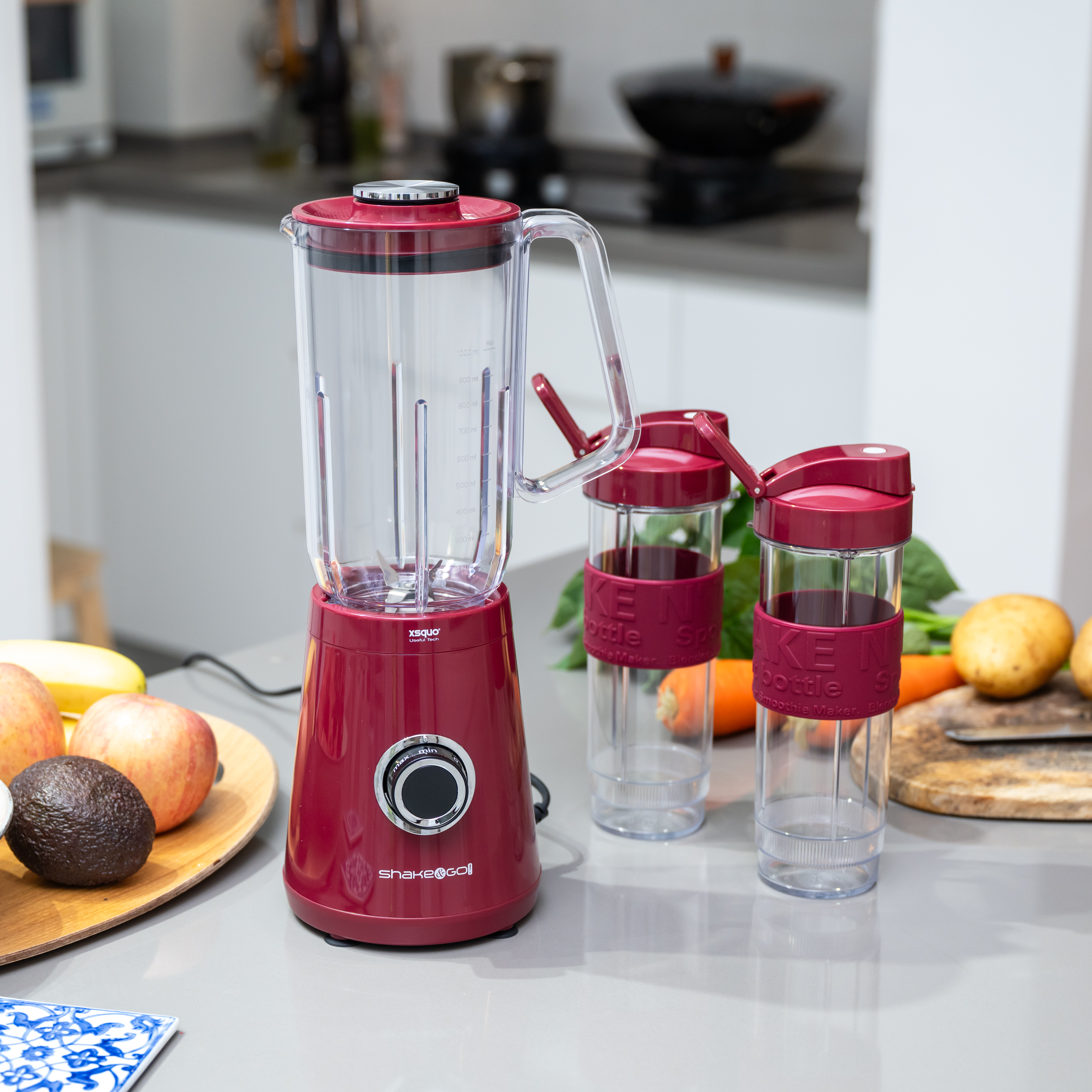 Una licuadora roja con dos botellas, fruta y verduras en una encimera de cocina. La licuadora está al lado de las botellas.