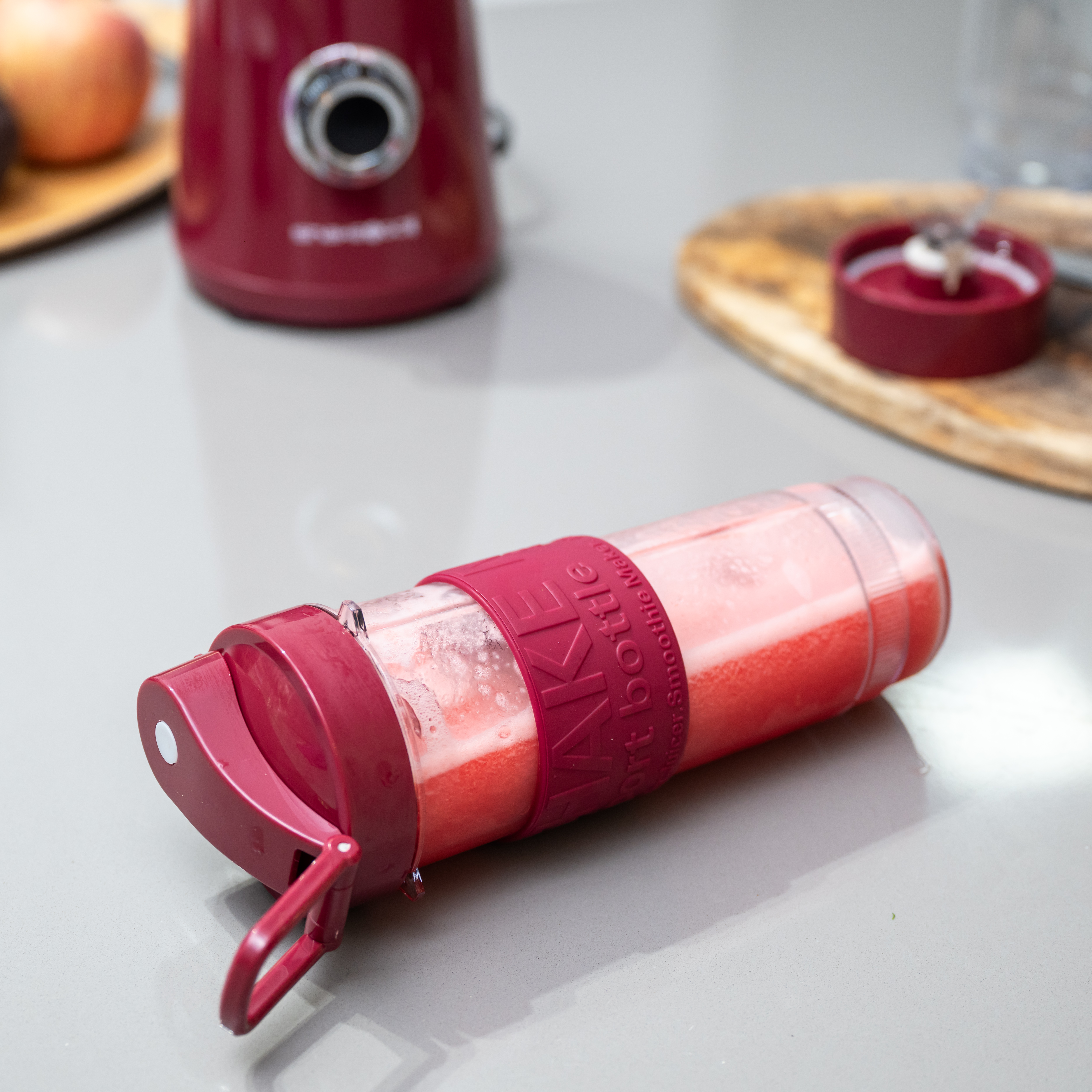 Una botella de batido roja con tapa y correa rojas sobre una superficie gris. Una licuadora y fruta en el fondo.