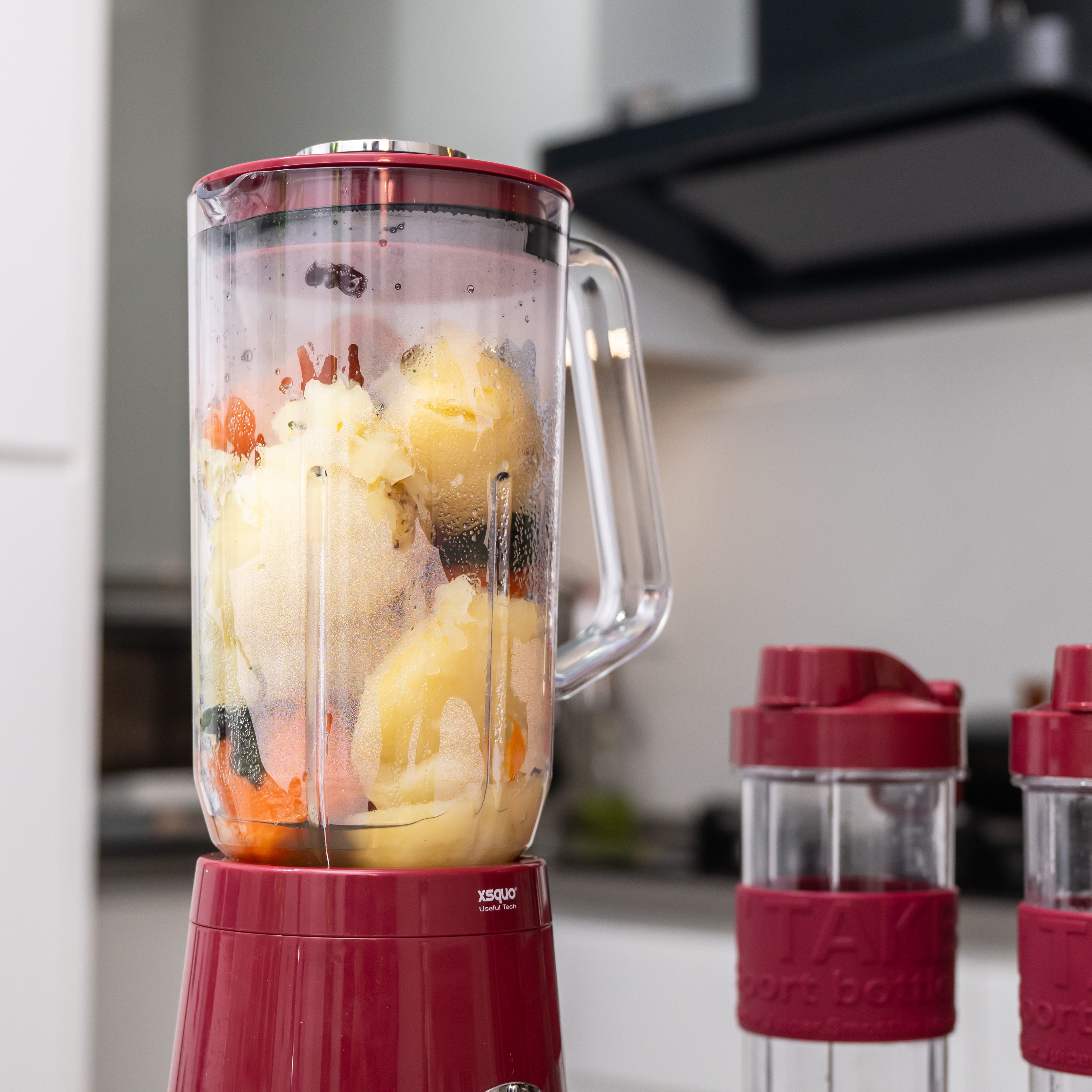 Una licuadora roja llena de alimentos amarillos y naranjas. Dos botellas deportivas a la derecha.