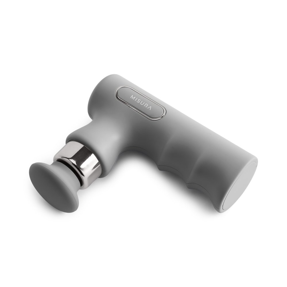 Szara pistolet do masażu Misura, ustawiona pod kątem w lewo, leży na białej powierzchni. Posiada głowicę do masażu, srebrne akcenty i uchwyt.