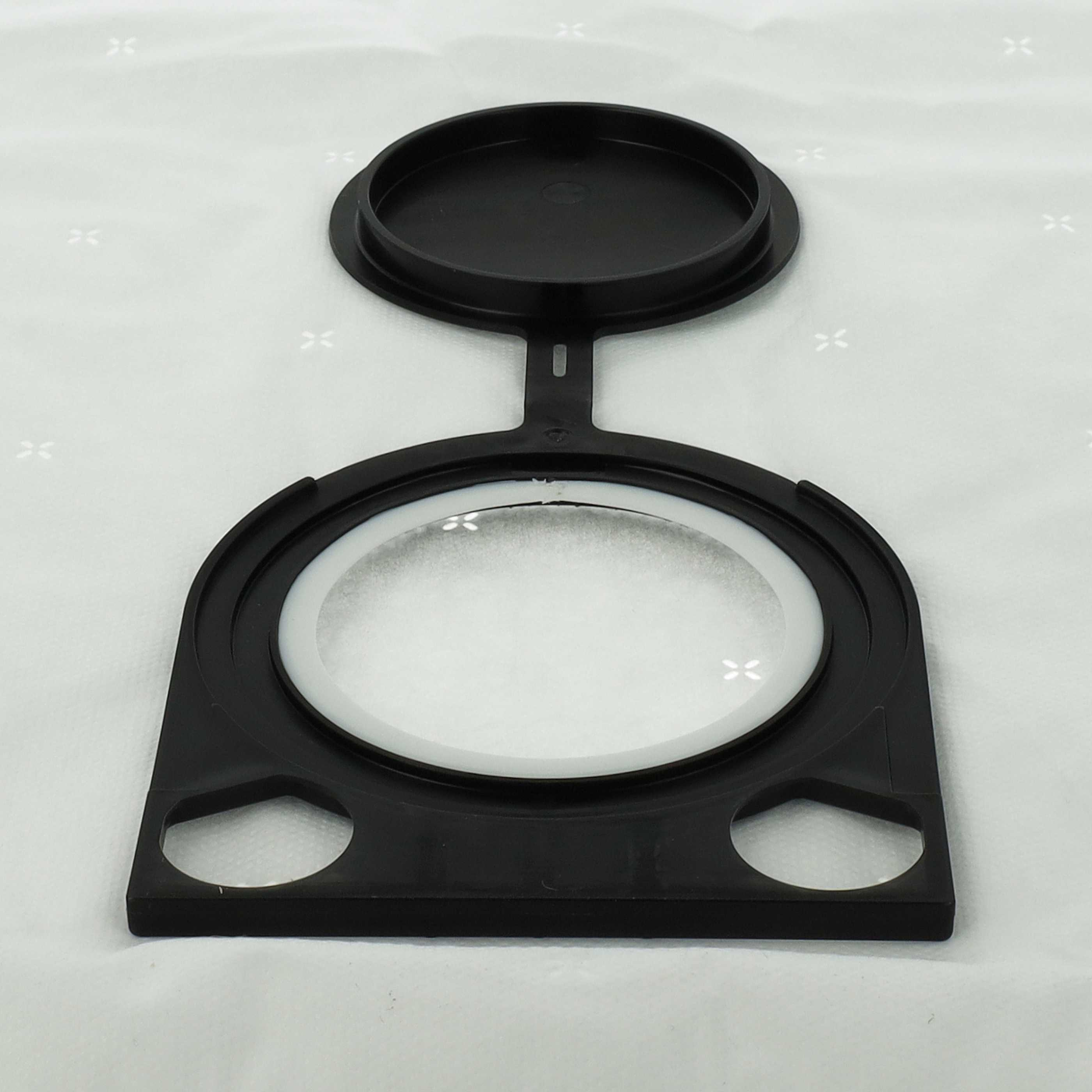 Ein schwarzes Kunststoffgerät mit runder Öffnung und Deckel liegt auf weißer Oberfläche. Das Gerät hat Linse und zwei Löcher.