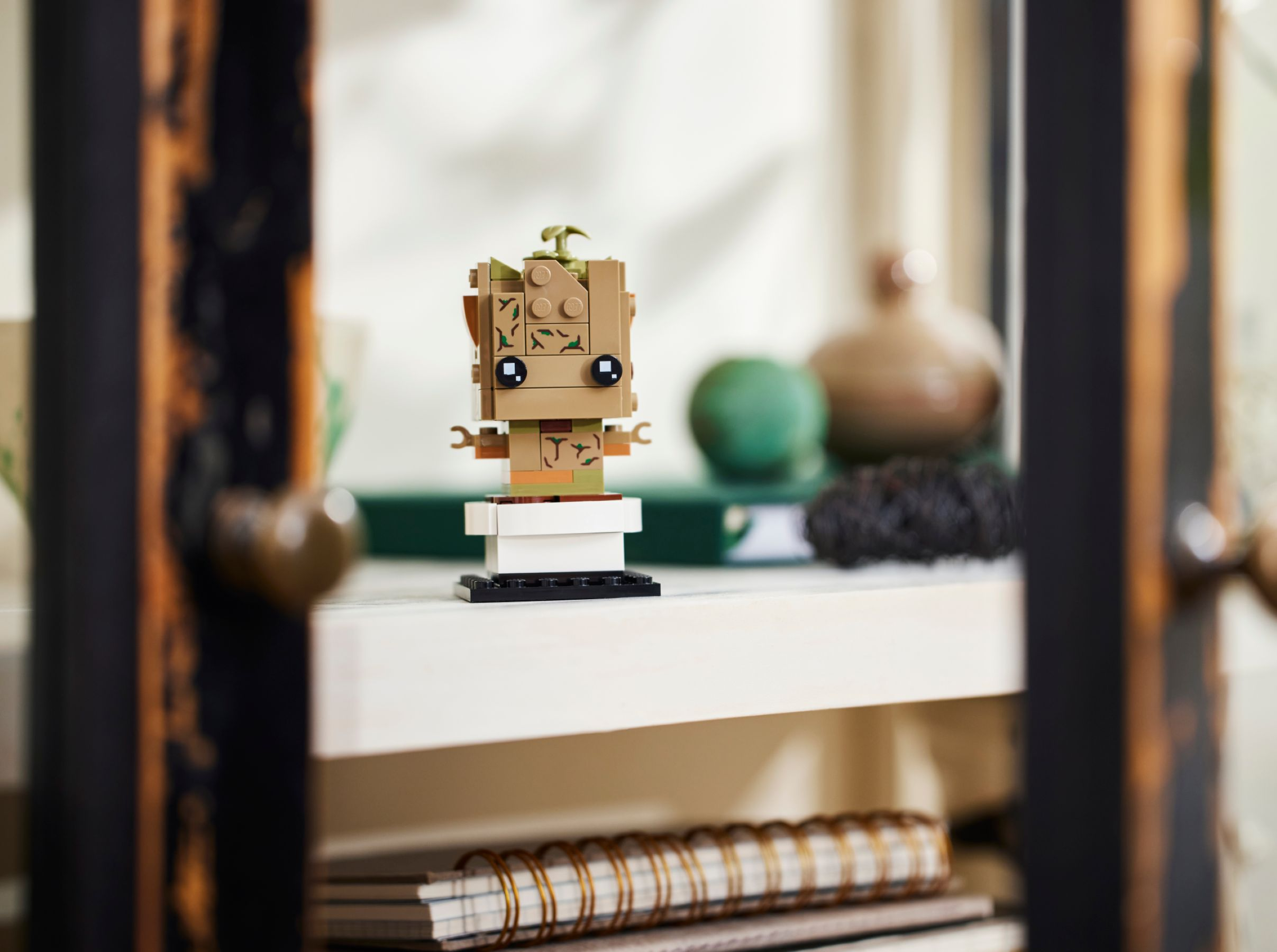 Lego Groot Figur auf einem Regal. Die Figur ist braun und grün, mit schwarzen Augen. Andere Objekte sind im Hintergrund unscharf.