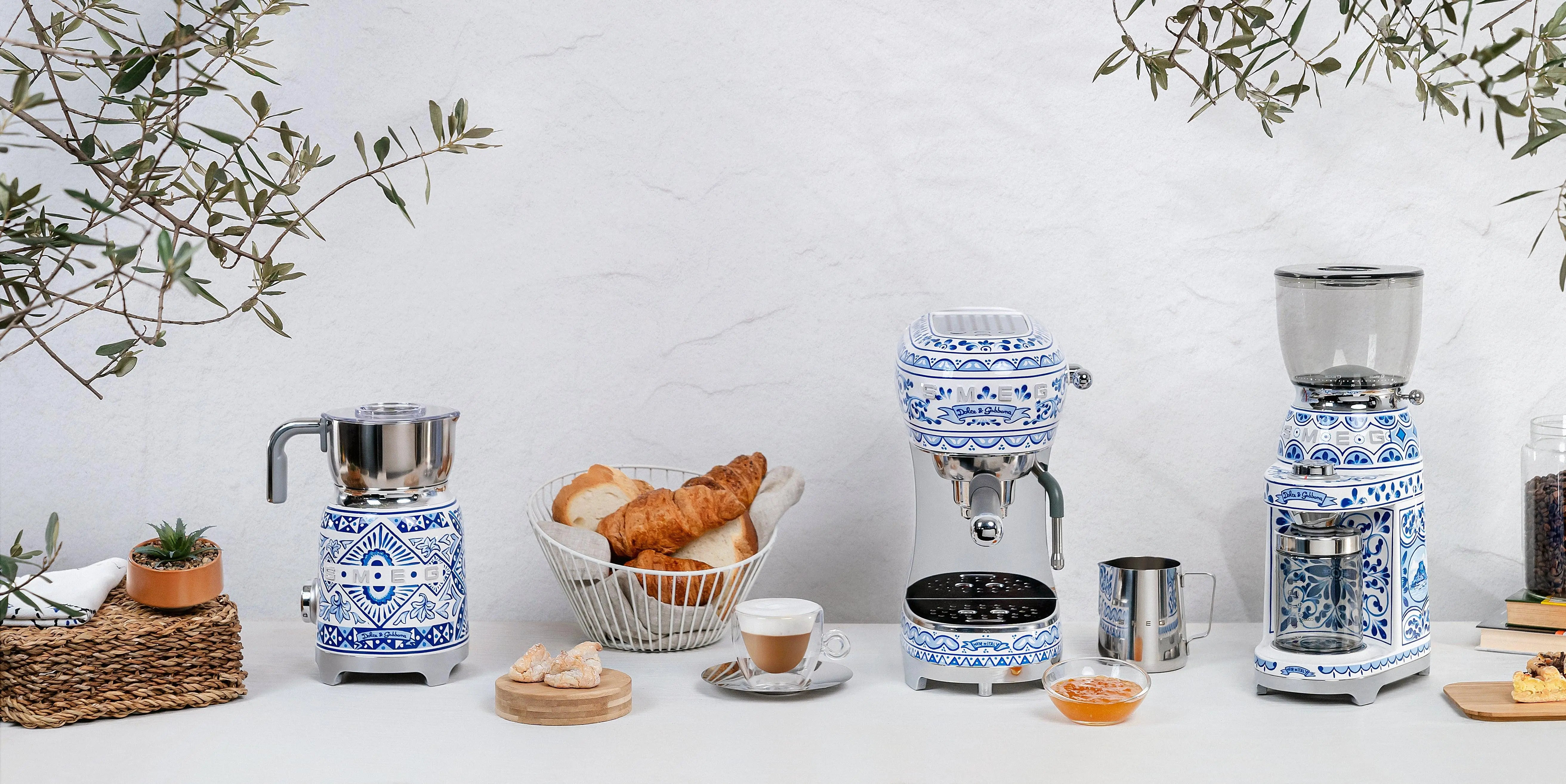 Eine Küchenszene mit Geräten und Lebensmitteln. Kaffeemaschine, Mühle, Gebäck und Kaffeetasse auf weißer Oberfläche, mit Olivenzweigen darüber.