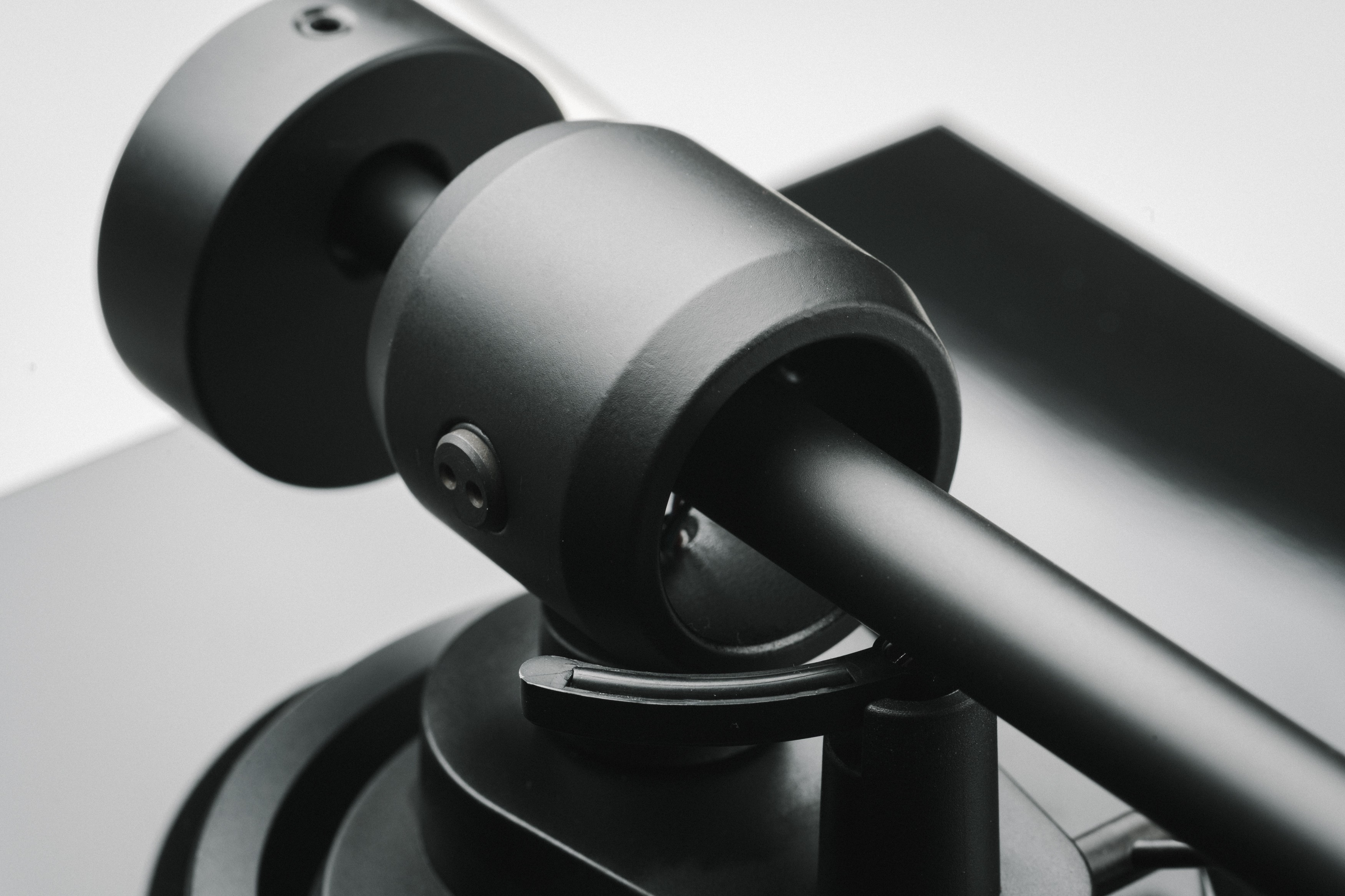 Zbliżenie czarnego ramienia gramofonu. Ramię jest cylindryczne, z zakrzywioną podstawą. Tło jest szare i białe.