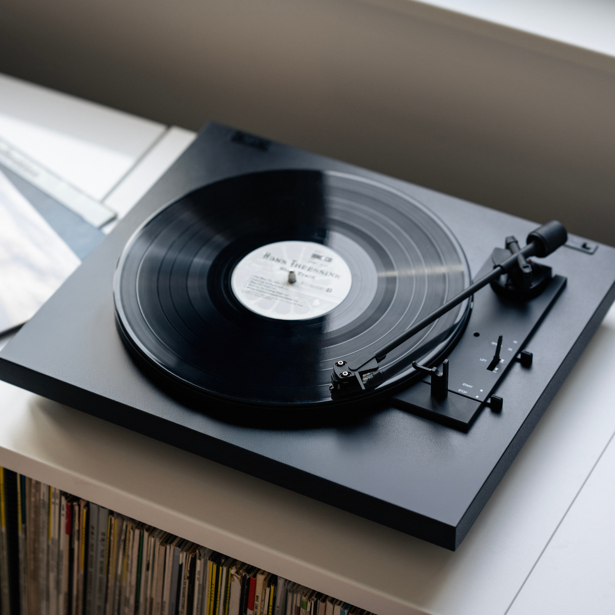 Een zwarte platenspeler met een vinylplaat staat op een wit oppervlak. Daaronder zijn vinylplaten gestapeld.