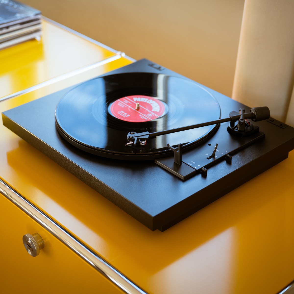 Een zwarte platenspeler met een vinylplaat op een gele tafel. Het label is rood en wit. Een stapel cd's op de achtergrond.