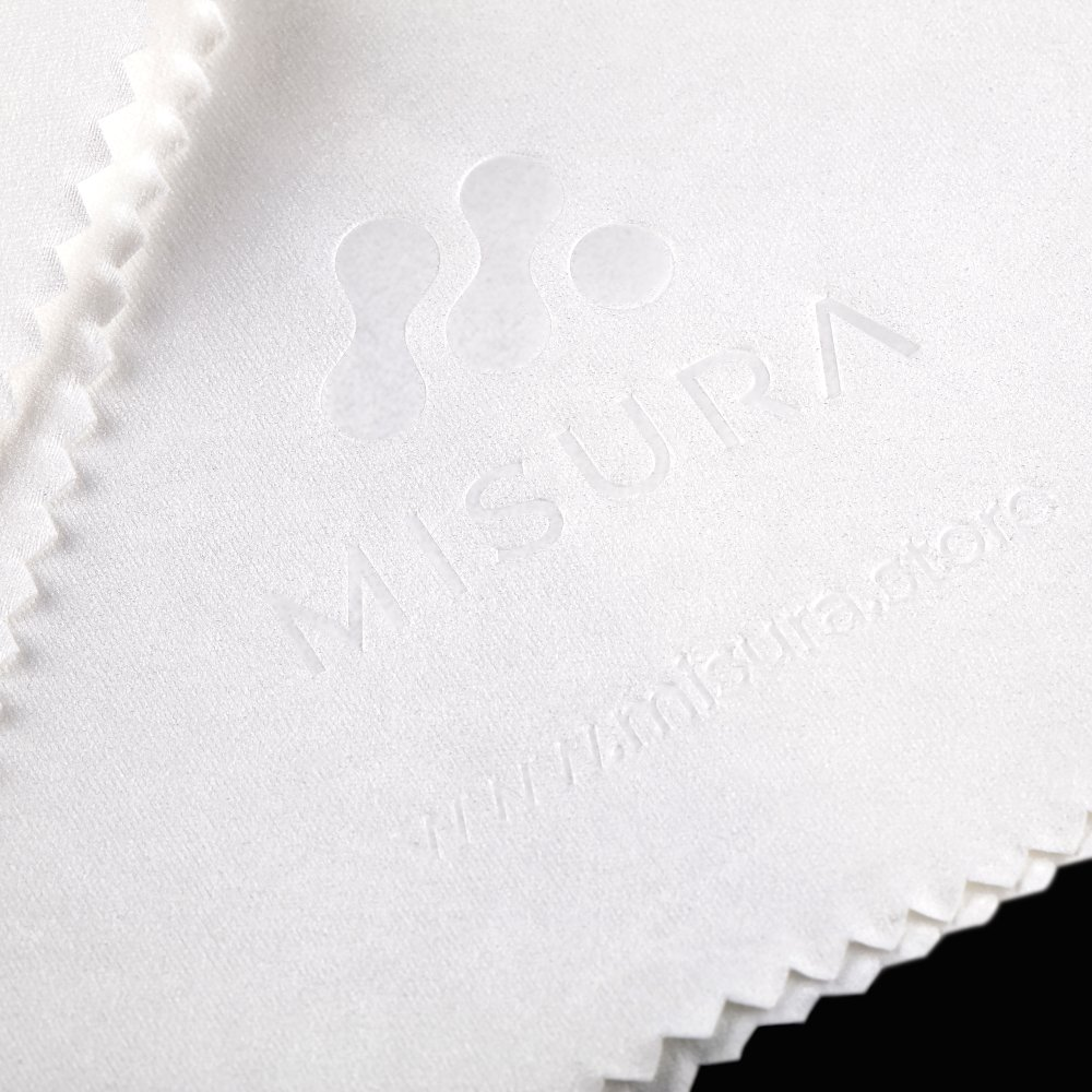 Paño de limpieza blanco con la palabra MISURA y la dirección del sitio web en relieve. El paño está sobre un fondo negro.