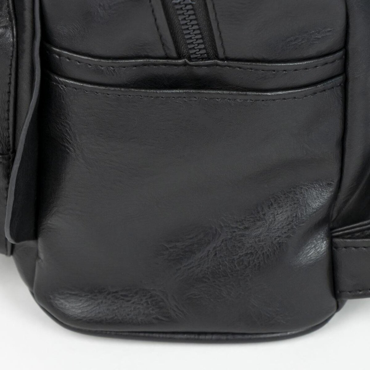 Primer plano de una bolsa de cuero negro con cremallera y bolsillo. El cuero es texturizado y brillante, sobre un fondo blanco.