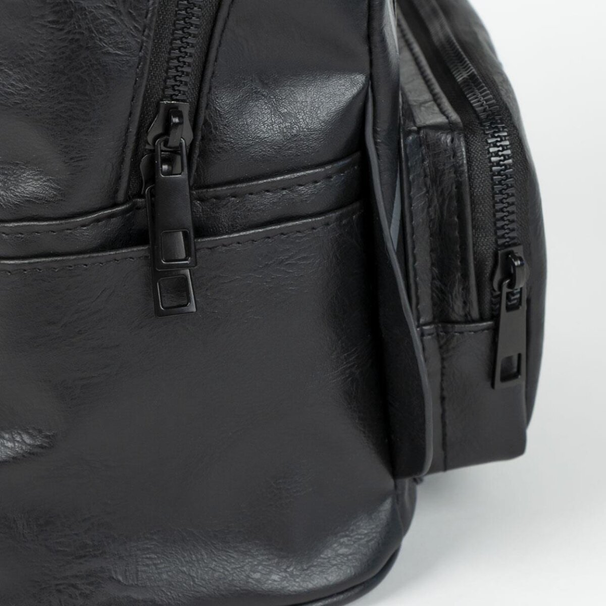 Primer plano de una bolsa de cuero negro. Se ven cremalleras, costuras y la textura del material sobre un fondo blanco.