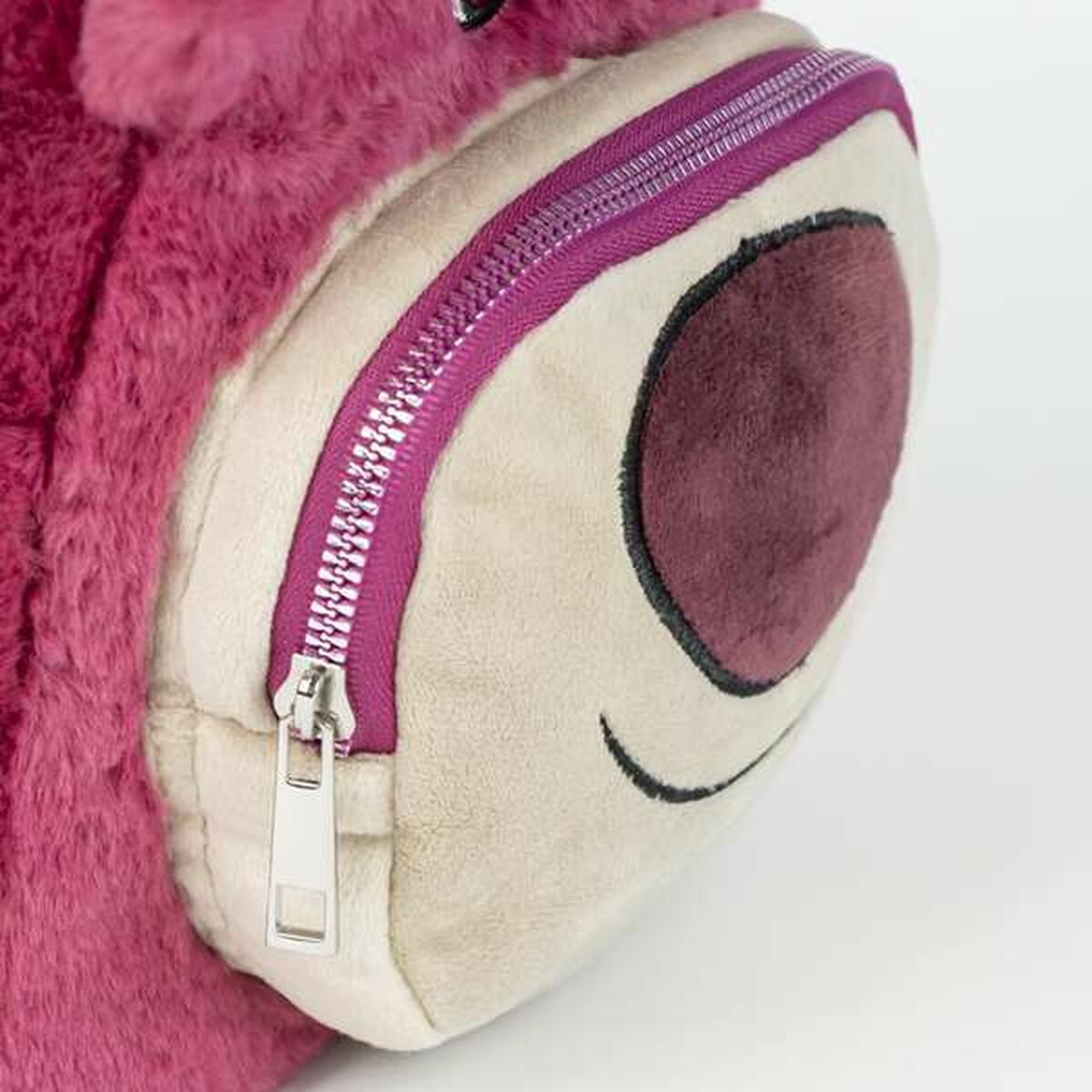 Primer plano de una mochila de peluche rosa con una cara color crema y una cremallera morada. El ojo es una forma ovalada de color púrpura oscuro.