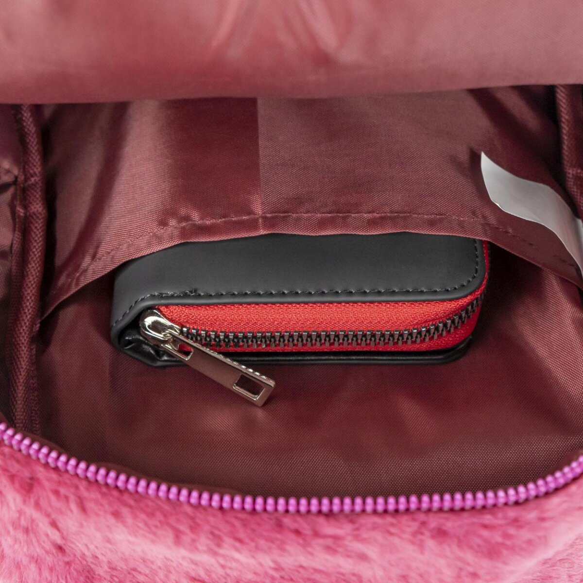 Dentro de una mochila rosa, se ve una cartera negra y roja con cremallera plateada. El fondo es de tela burdeos.