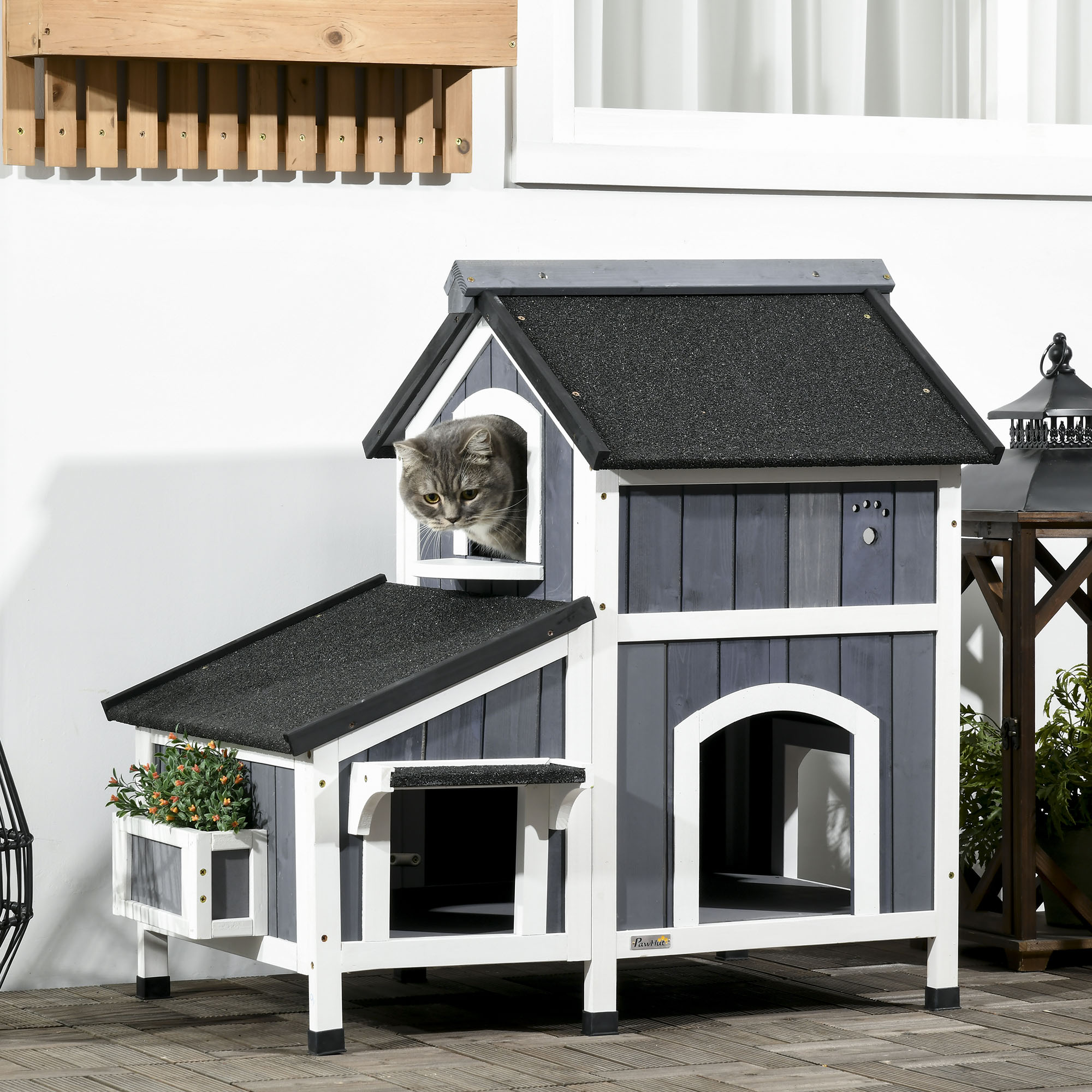 Un gato gris mira por una caseta para gatos gris y blanca. La casa tiene un tejado negro y una pequeña jardinera.