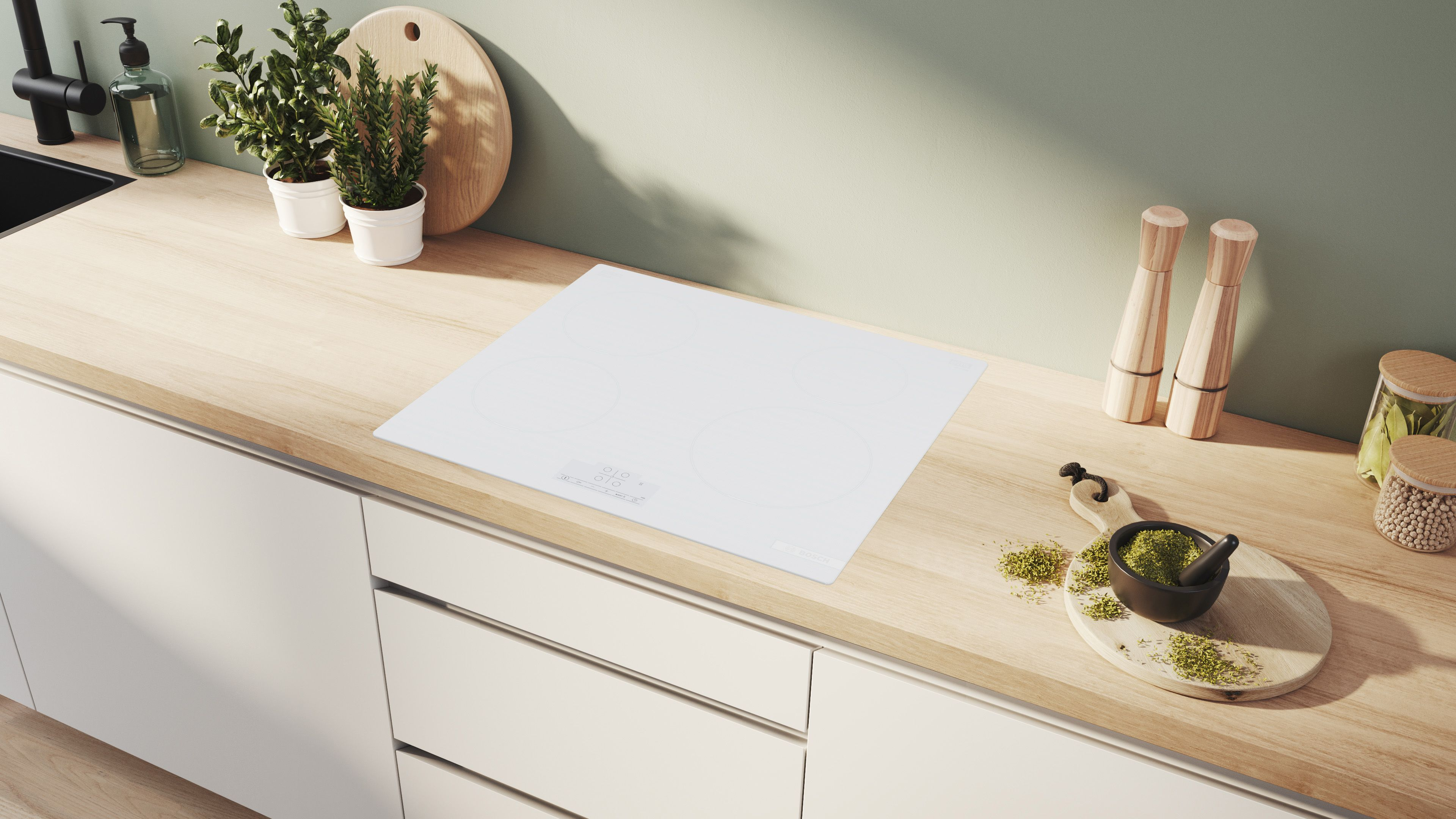 Scena kuchenna z białą płytą grzewczą, drewnianym blatem, roślinami i przyborami kuchennymi. Mędrzec i tłuczek na desce.
