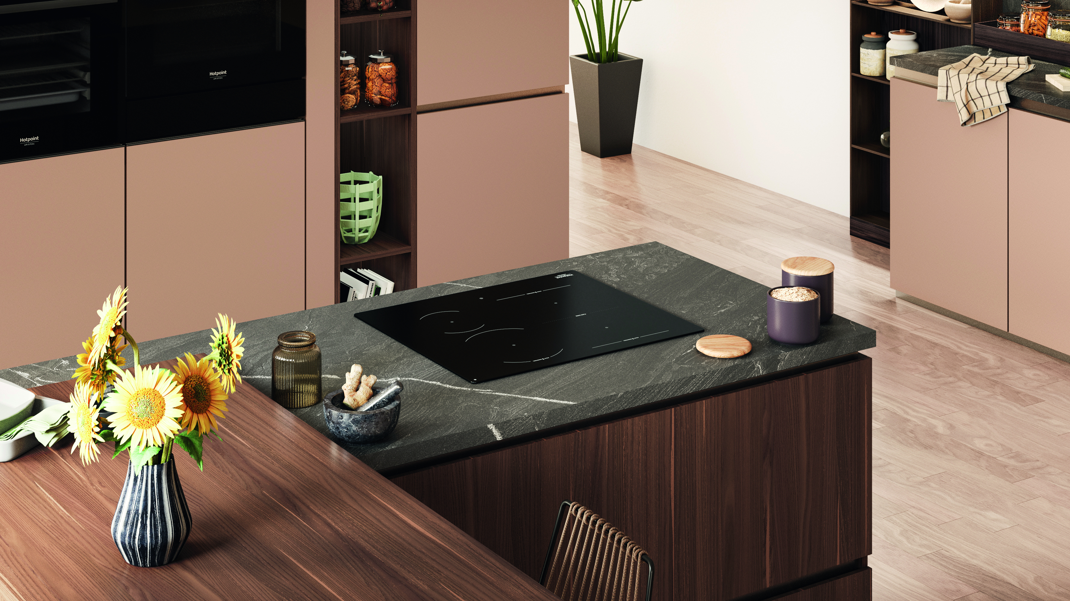 Una scena di cucina con un piano cottura nero, girasoli in un vaso e armadi rosa. Pavimento in legno e piano di lavoro scuro.