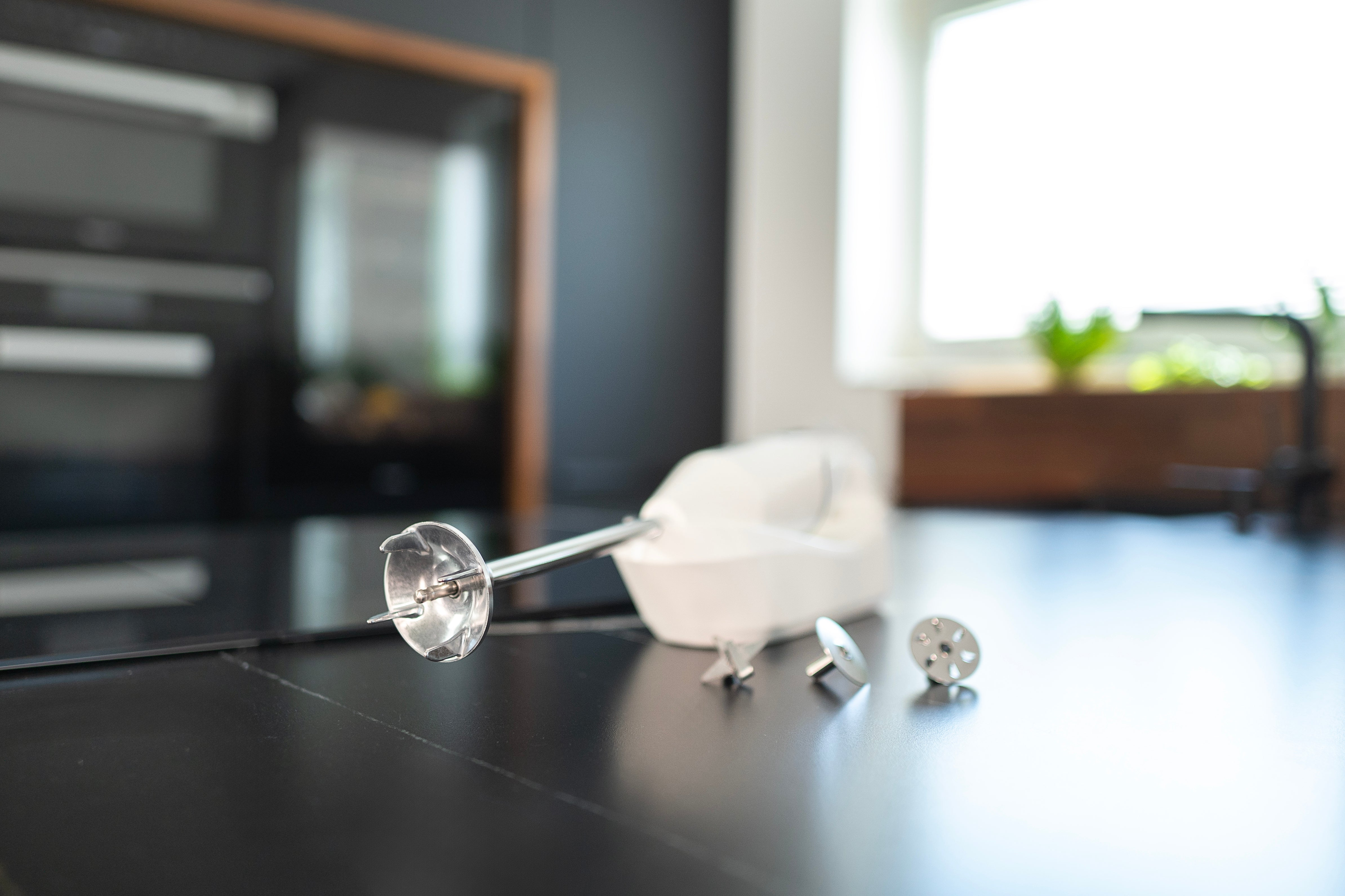 Ein weißer Handmixer mit Aufsätzen auf einer schwarzen Arbeitsplatte. Küchenhintergrund mit Backofen und Fenster.