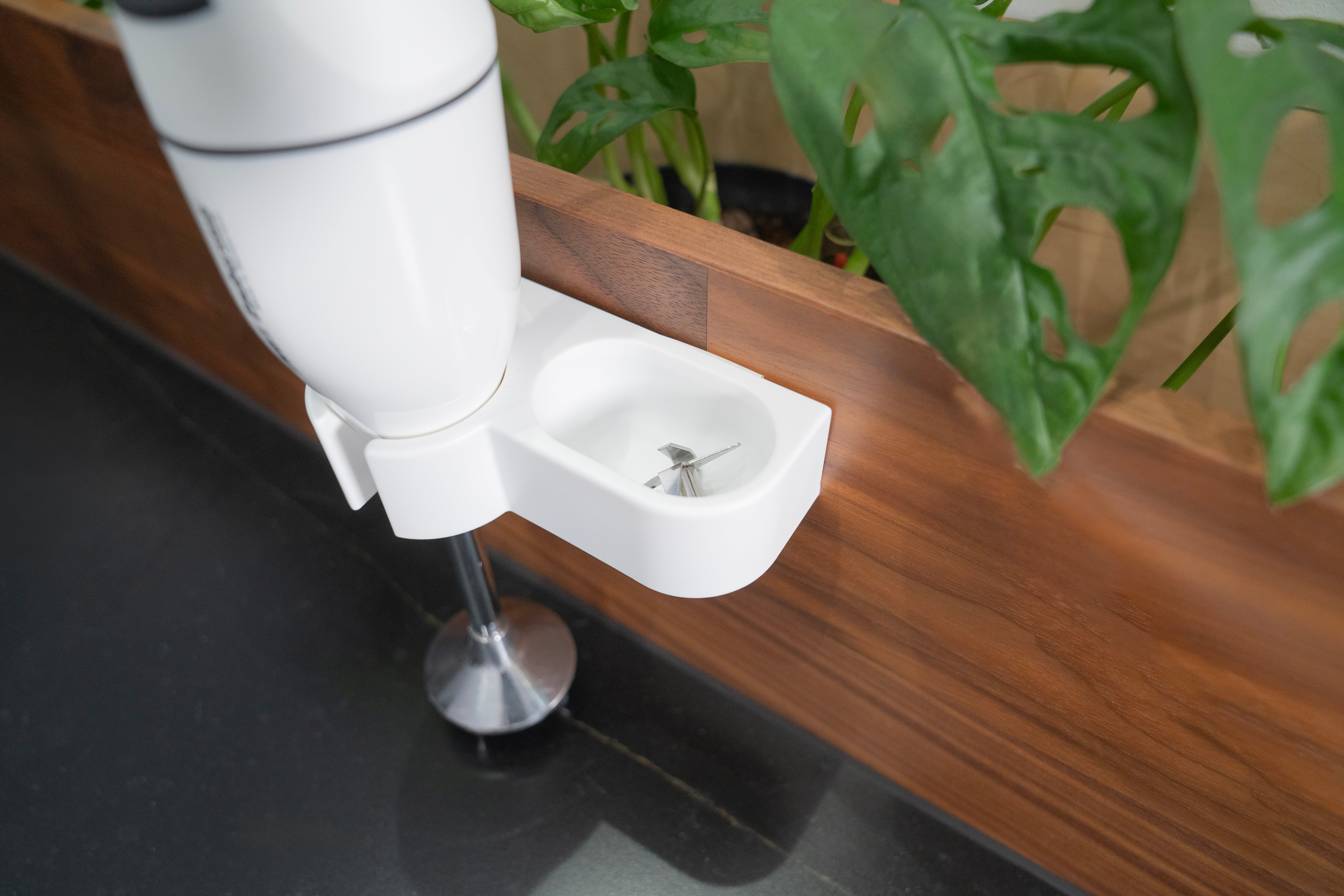 Ein weißer Stabmixer wird in einer Halterung platziert. Grünpflanze im Hintergrund und dunkle Arbeitsplatte.