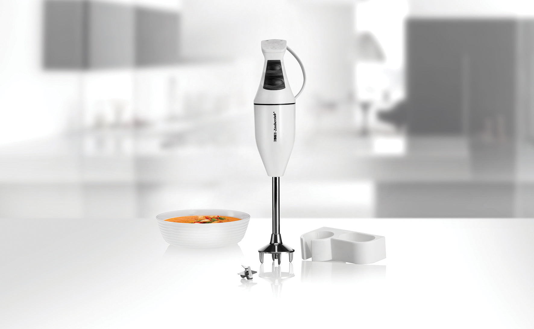 Biały blender ręczny z akcesoriami stoi obok miski z zupą na białej powierzchni. Tło to rozmyta kuchnia.