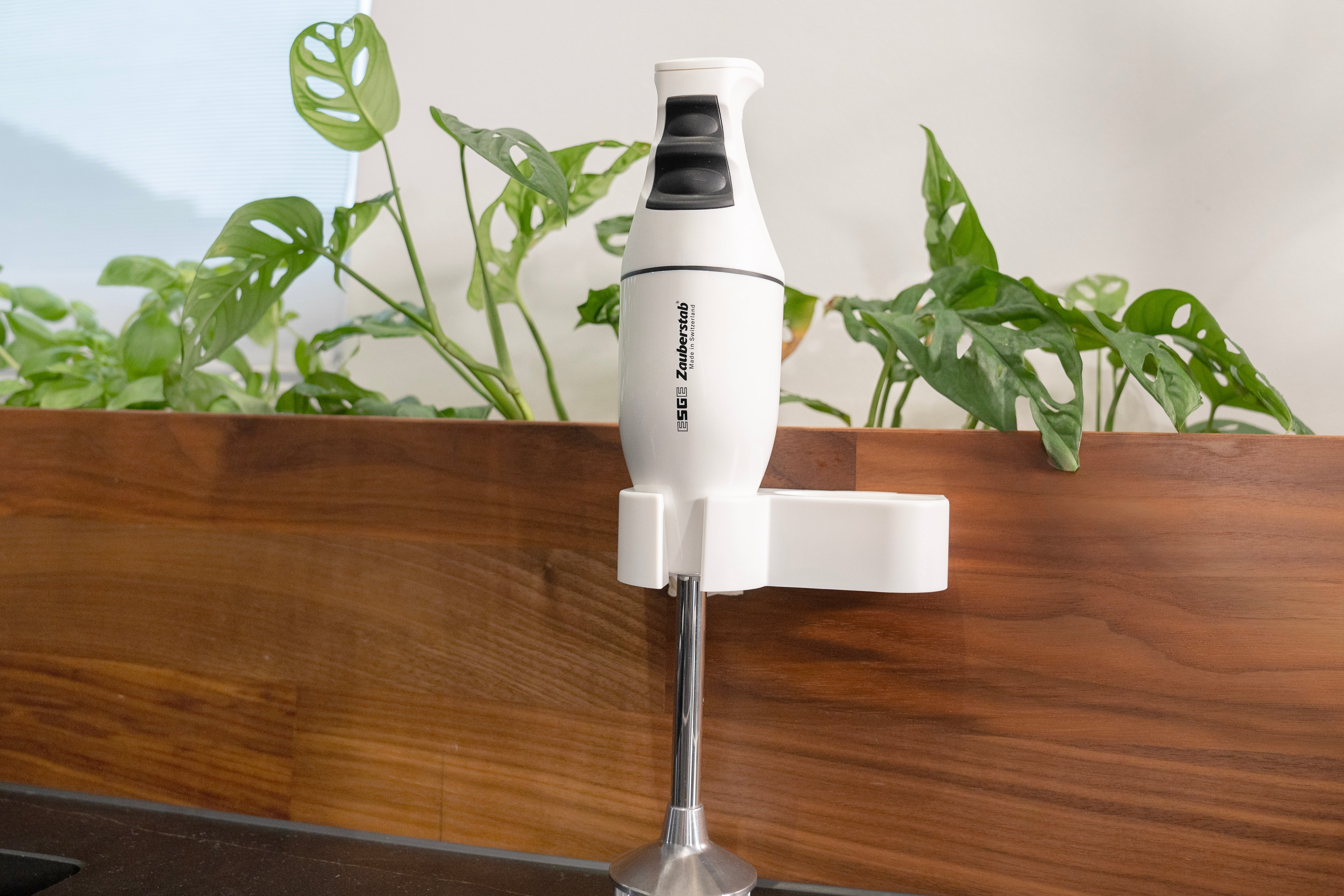 Biały blender ręczny jest zamontowany na uchwycie. Zielone rośliny w drewnianej donicy. Tło: ściana i okno.