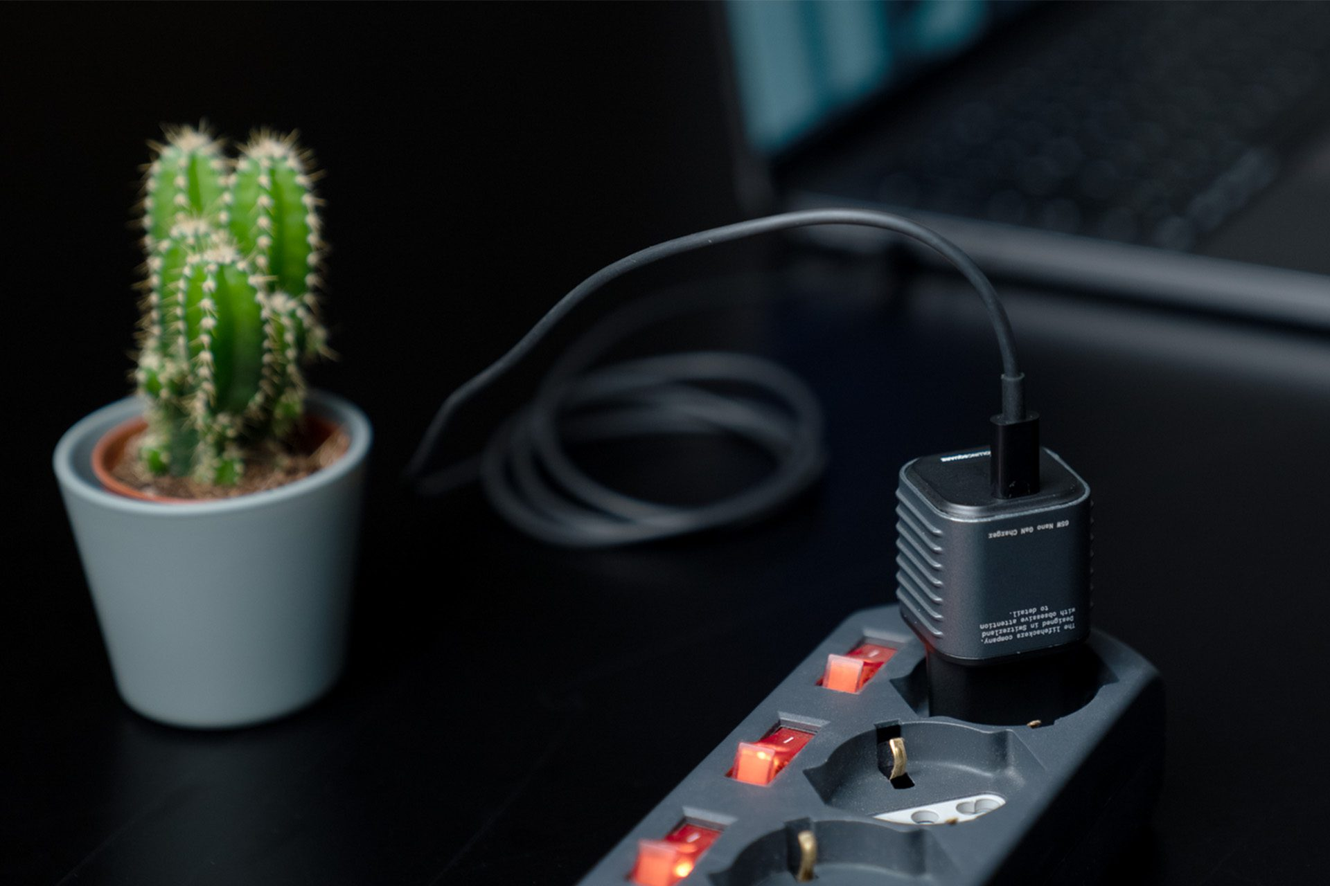 A cactus in a pot, charger plugged into a power strip. Laptop in the background. Focus on the charger and cactus.