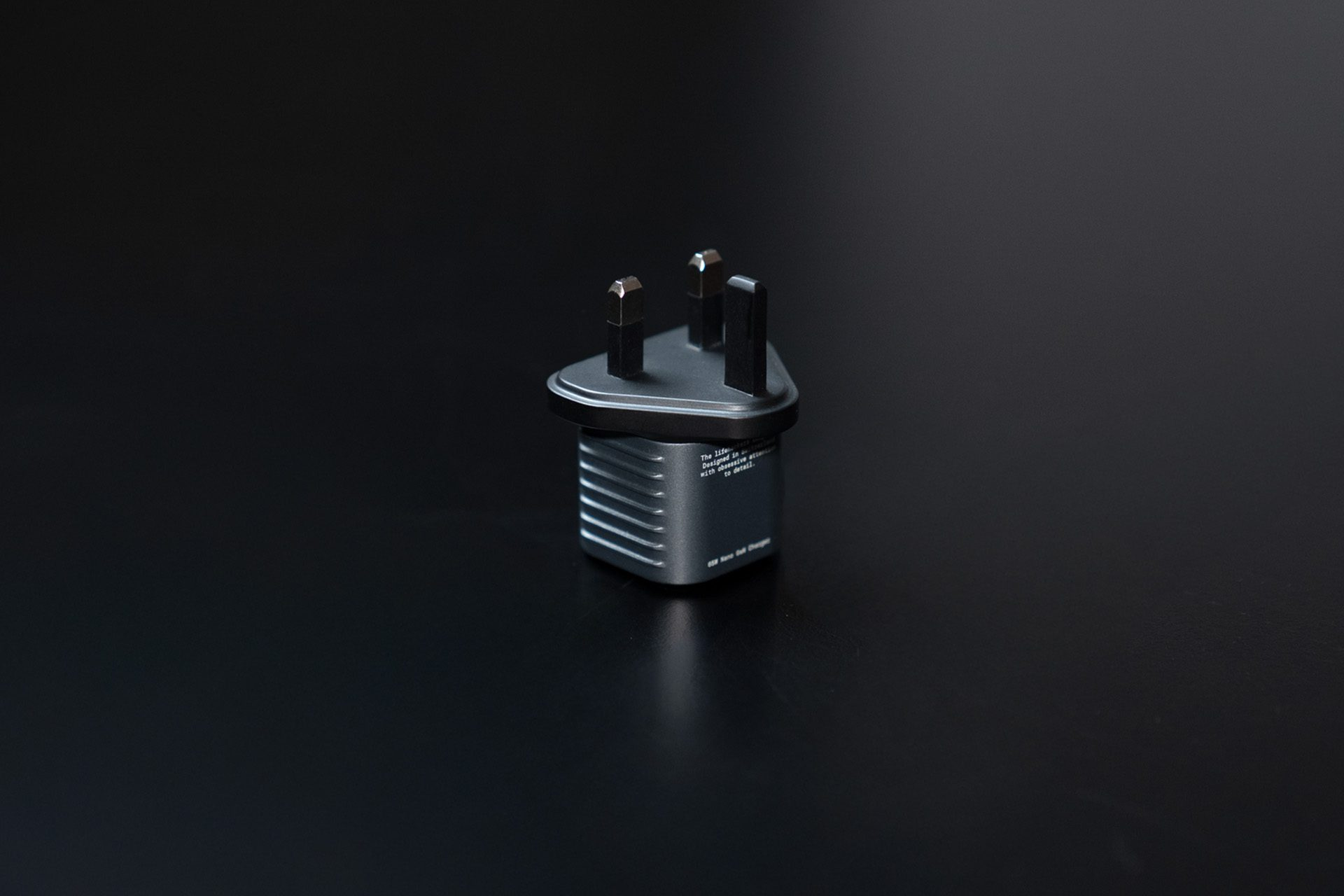 A gray UK plug adapter with three prongs on a black surface. The adapter is the main subject, well-lit against the dark background.