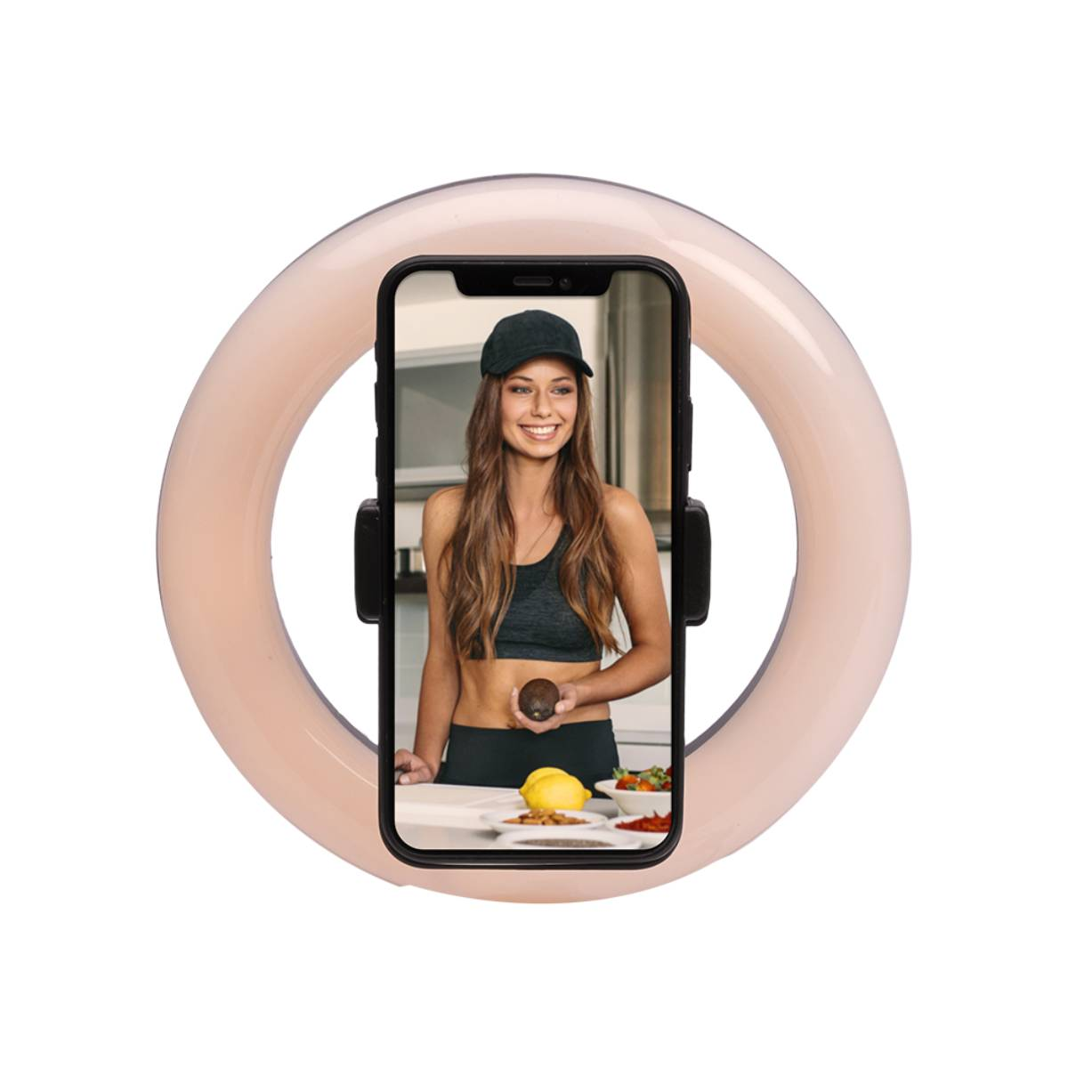 Femme en casquette noire et soutien-gorge de sport tenant un avocat devant un téléphone dans un anneau lumineux. Cuisine avec nourriture.