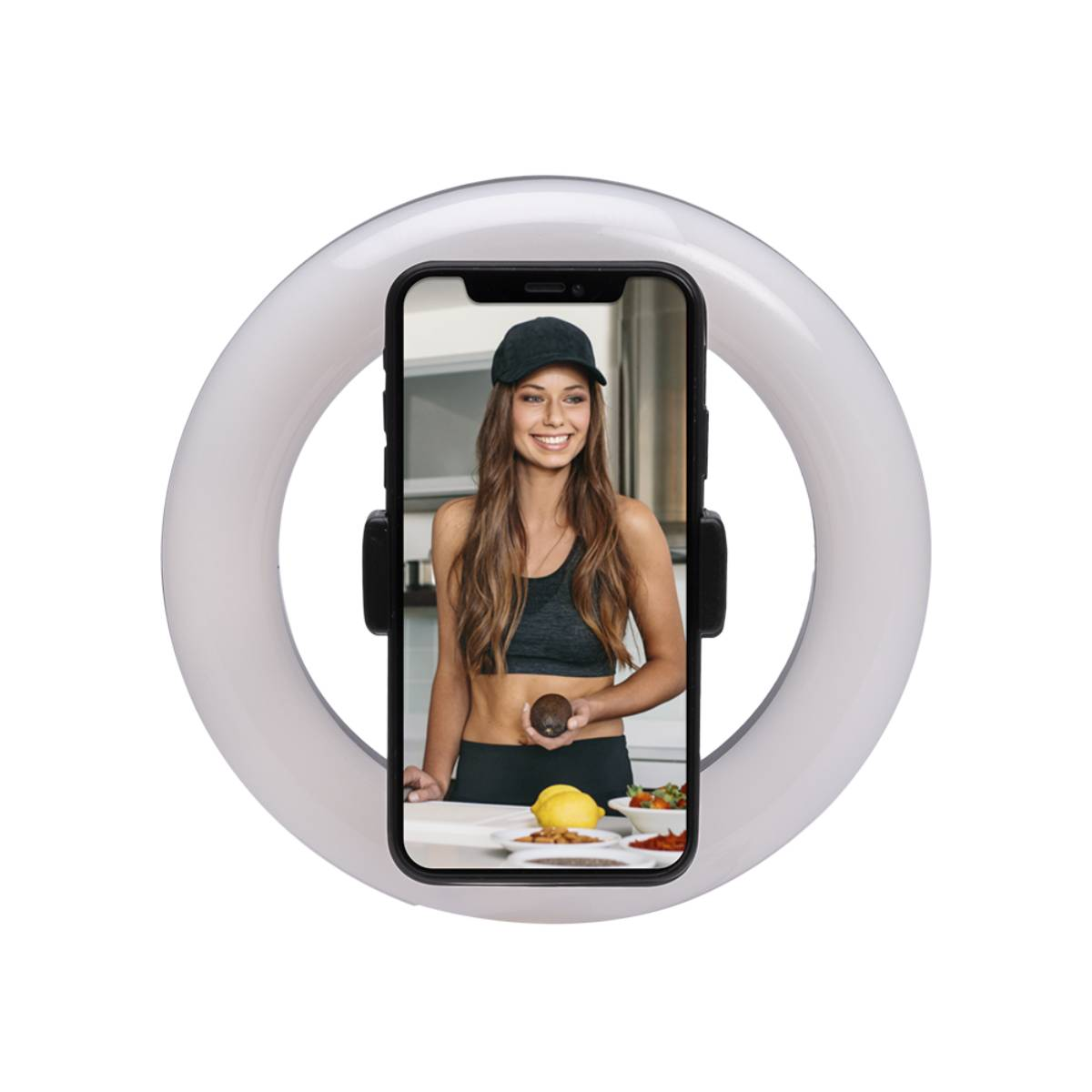 Une femme tient un avocat devant une caméra, à l'intérieur d'un anneau lumineux. Elle porte une casquette et un soutien-gorge de sport, avec de la nourriture sur le comptoir.
