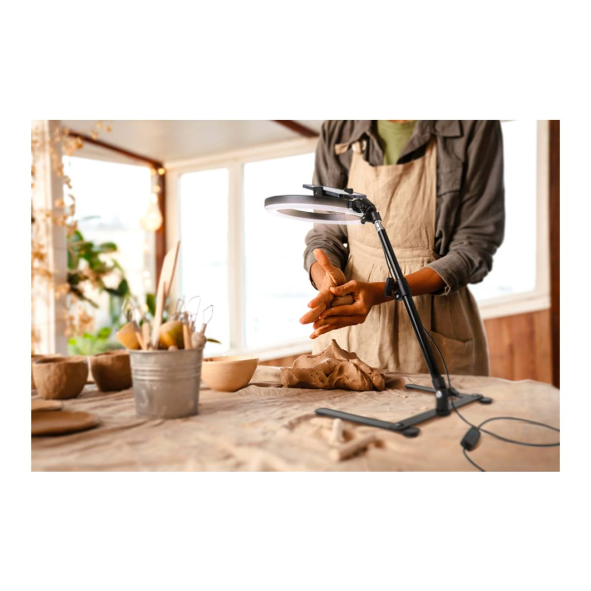 Une personne travaille l'argile sur une table. Une lampe éclaire la scène. Des outils de poterie, des bols et de l'argile sont sur la table.