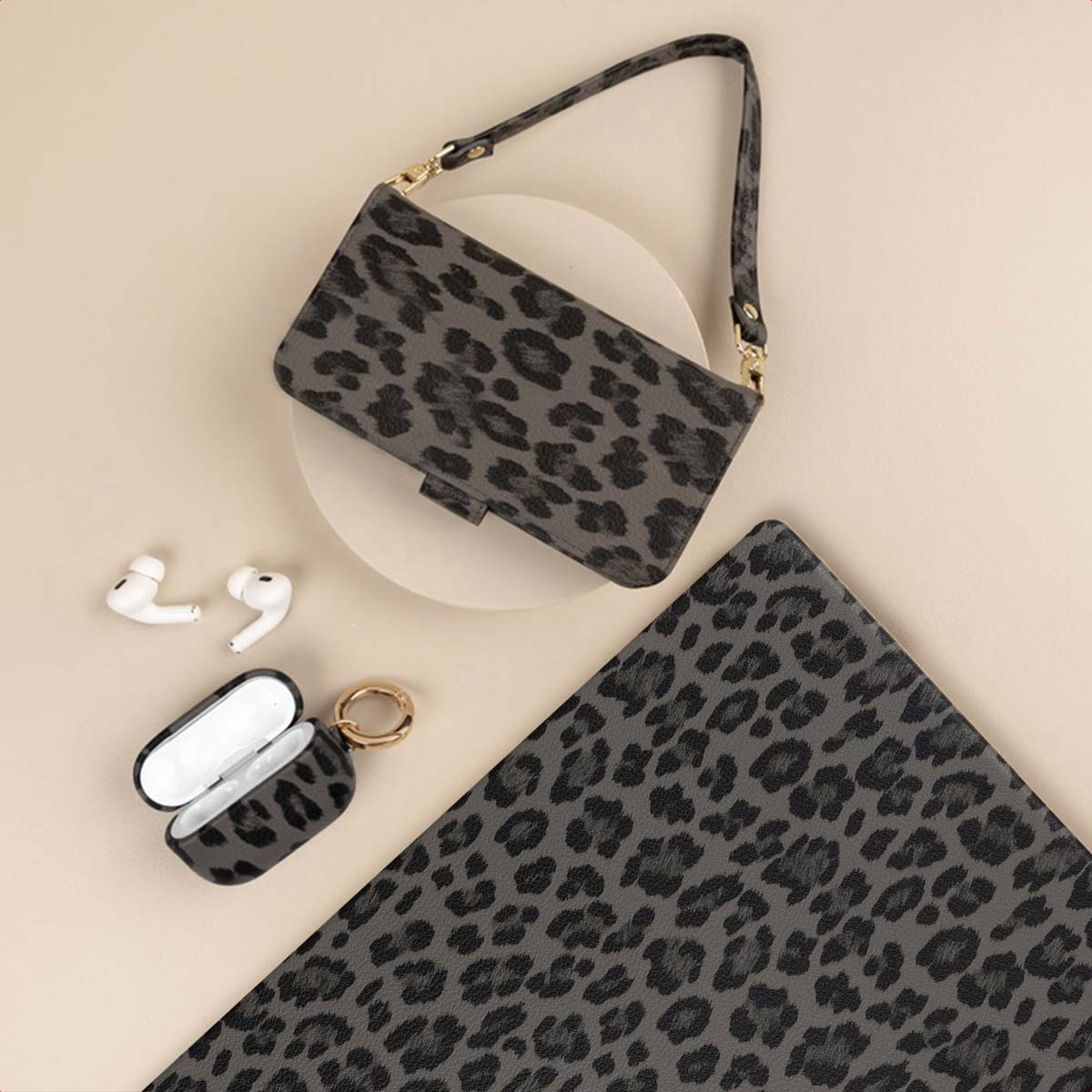Un sac à main gris à imprimé léopard, un étui de téléphone et un étui AirPods sont disposés sur une surface beige, à côté d'AirPods blancs.