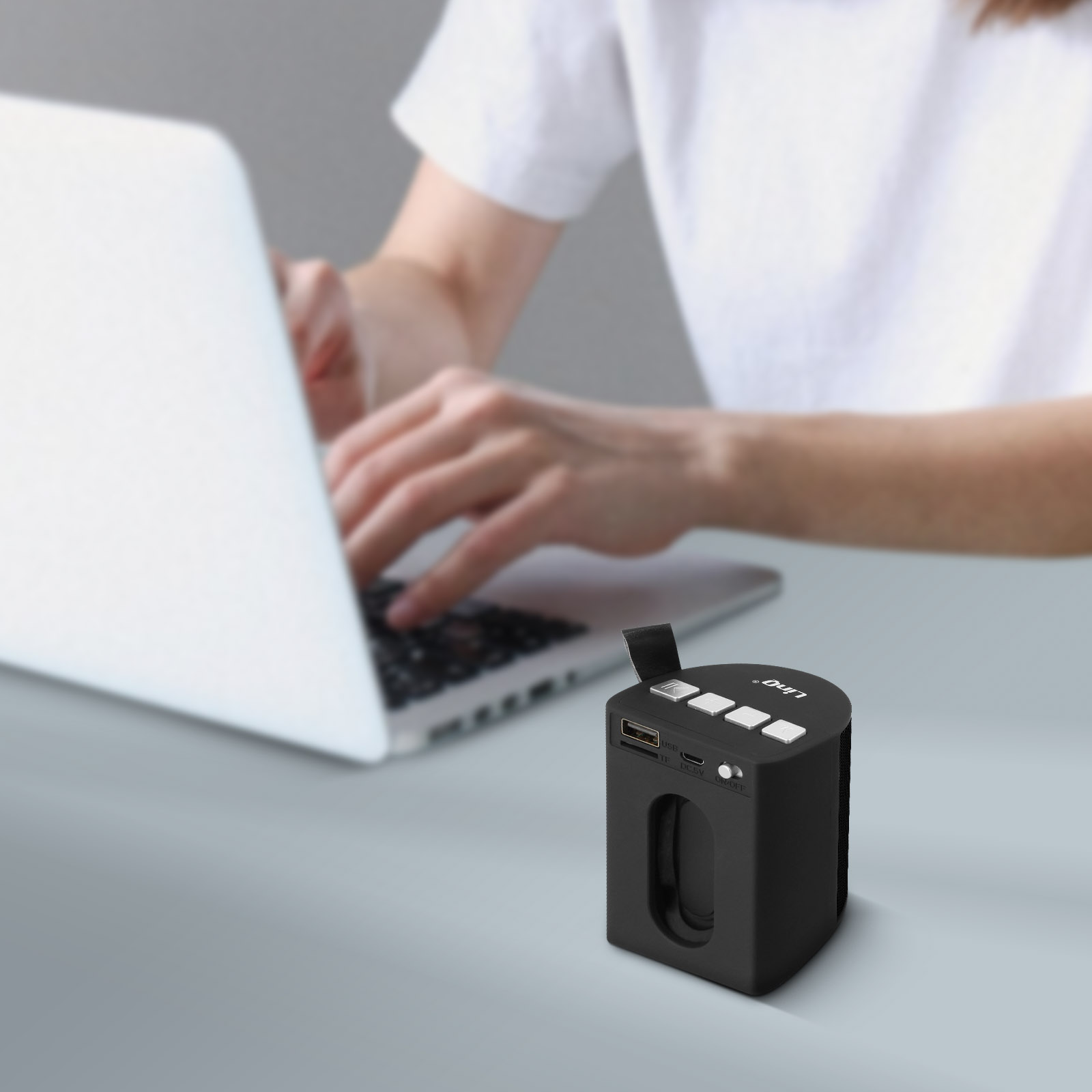 Una persona usa una computadora portátil. Un altavoz negro está en la mesa. El altavoz tiene botones y un puerto USB.