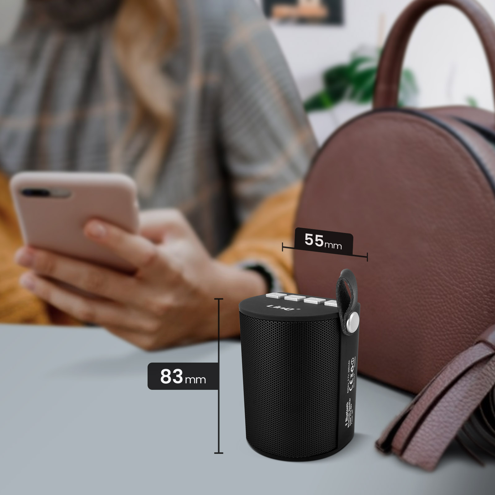 Un altavoz negro de 55x83 mm, una mujer con un teléfono y una bolsa marrón al fondo.