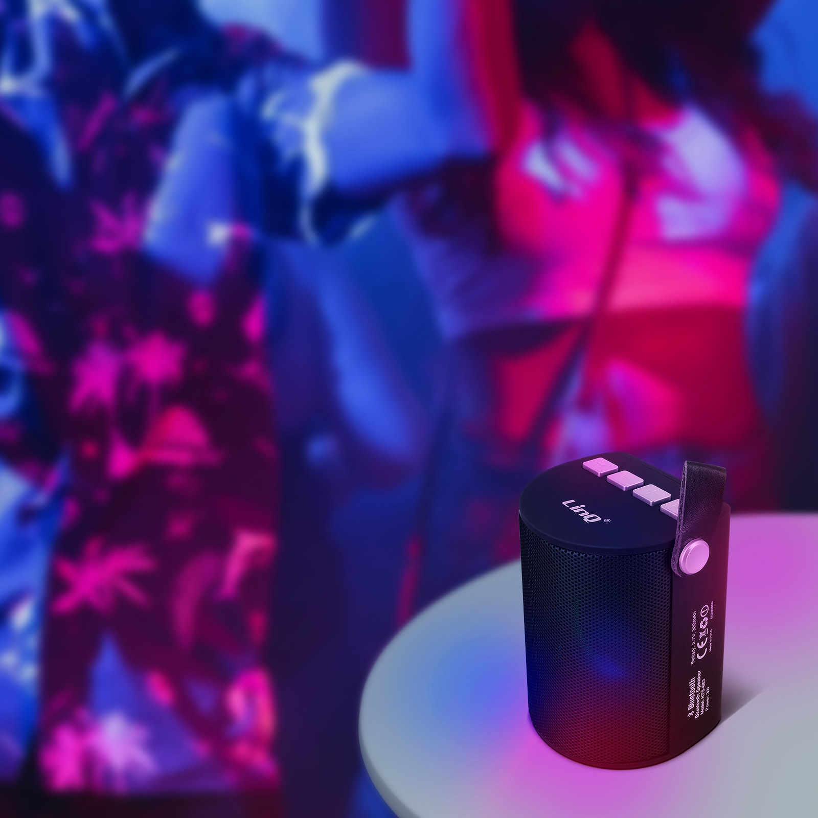 Un altavoz negro con botones está sobre una mesa, con personas borrosas en el fondo. Luces rosas y azules iluminan la escena.