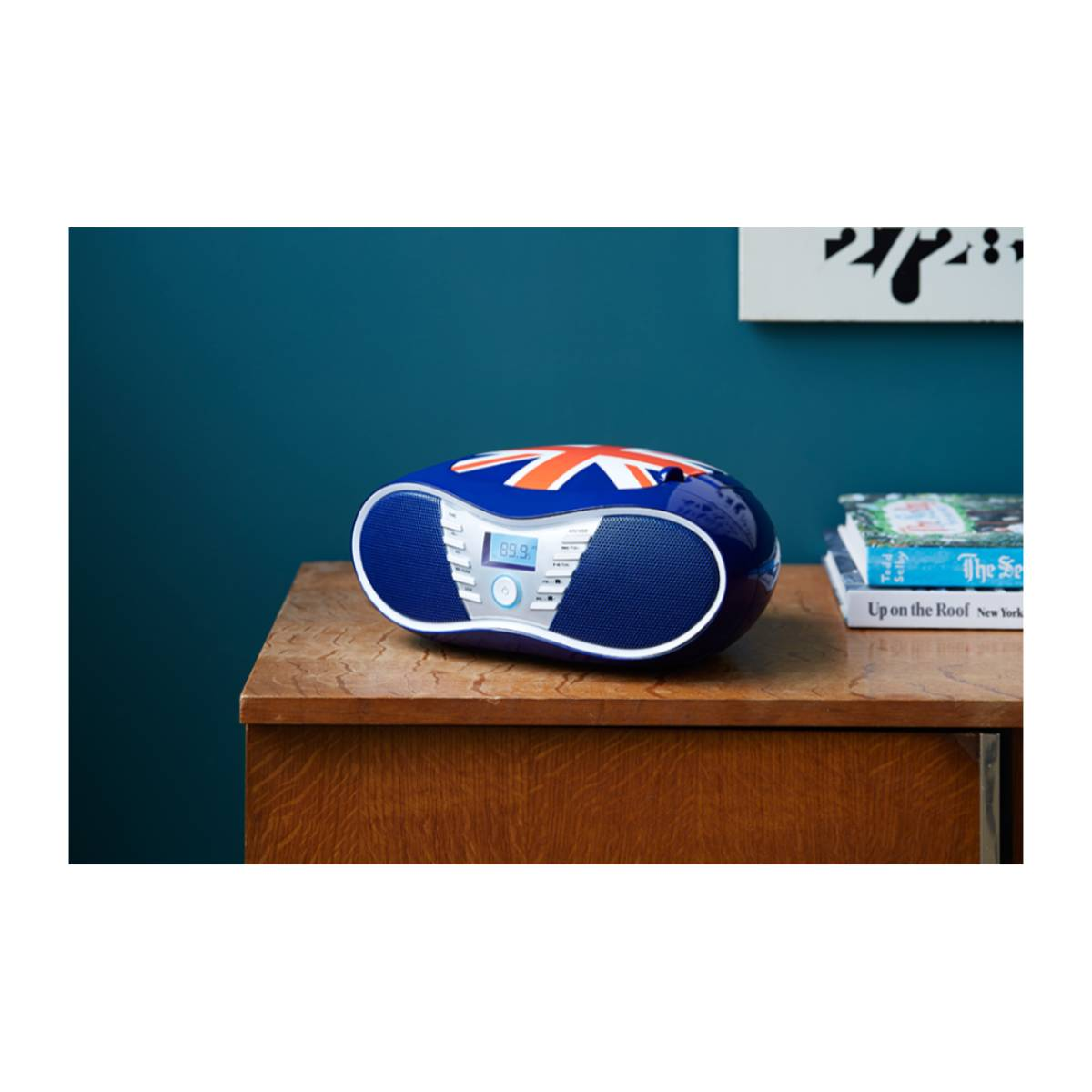 Una radio blu con un design Union Jack è su una superficie di legno. Libri e un dipinto sullo sfondo contro un muro verde acqua.