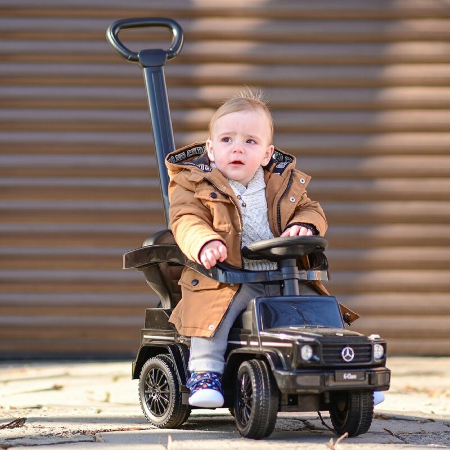 Ein Baby sitzt in einem schwarzen Spielzeugauto mit Schiebegriff. Das Baby trägt eine braune Jacke und graue Hose. Dahinter eine Holzwand.