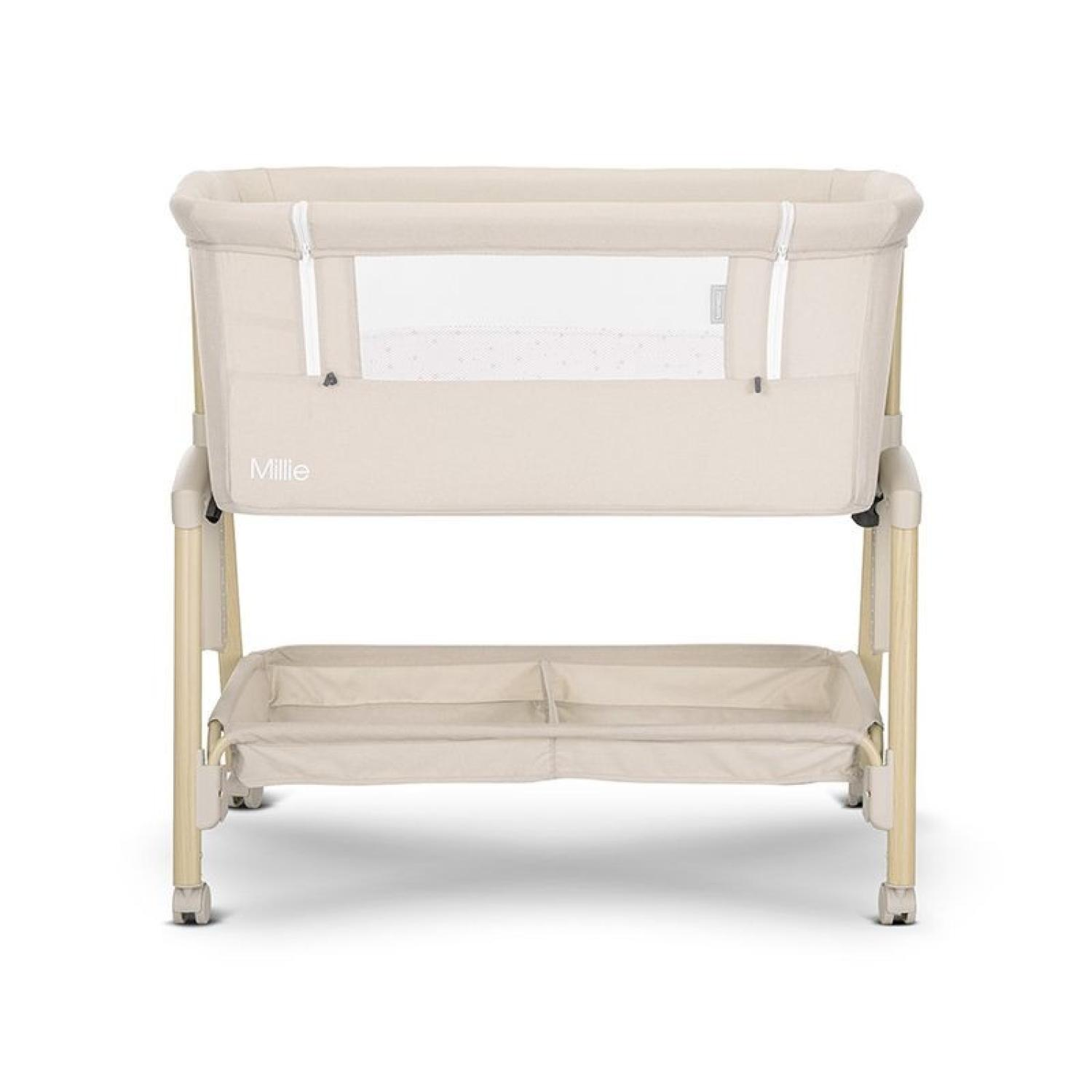 Una cuna beige con un lateral de malla, patas de madera y un estante de almacenamiento. Se ve el nombre "Millie".