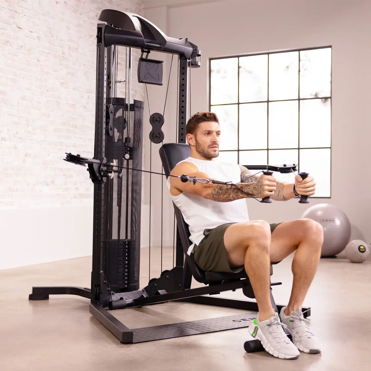 Mann benutzt eine schwarze Gewichtsmaschine und zieht Griffe zur Brust. Er trägt ein weißes Oberteil und Shorts, mit Tattoos. Fitnessstudio.