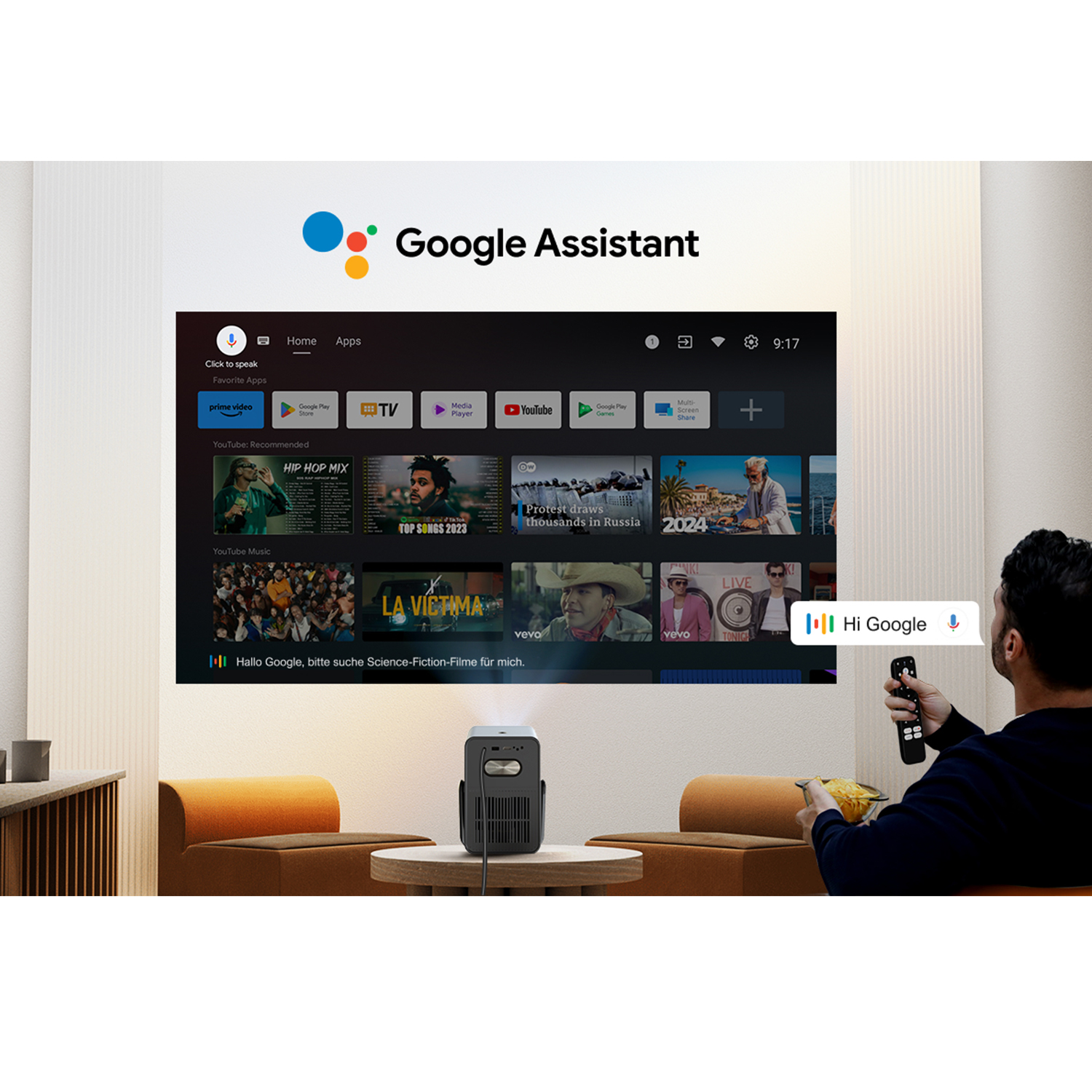 Una persona usa un telecomando con un proiettore che mostra un'interfaccia TV. Google Assistant è sullo schermo. Un piccolo proiettore è su un tavolo.