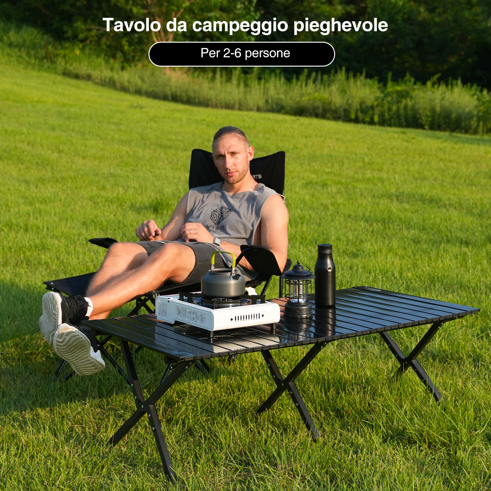 Un uomo è seduto su una sedia pieghevole a un tavolo nero sull'erba. Un fornello da campeggio, un bollitore, una lanterna e una bottiglia sono sul tavolo.
