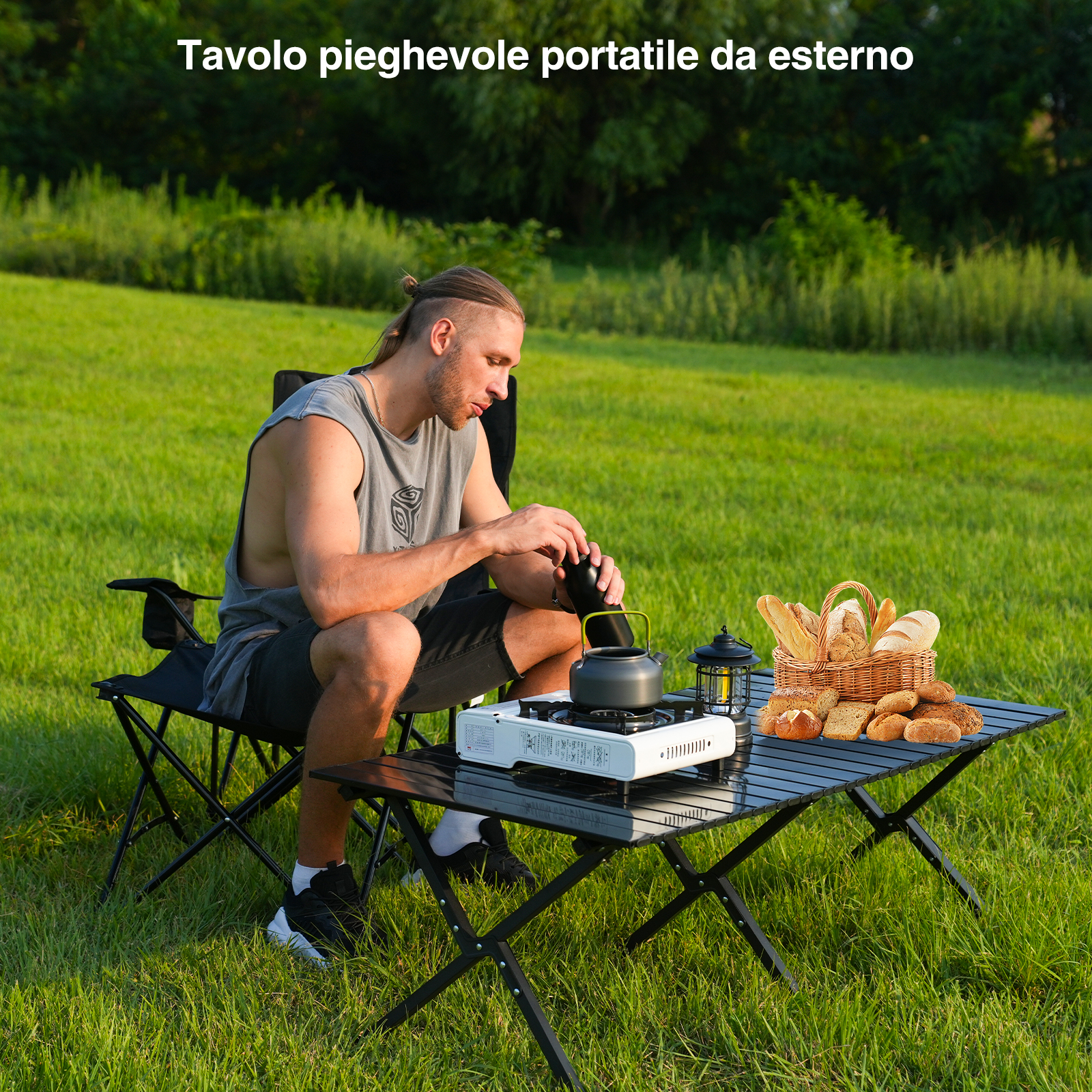 Uomo su una sedia, seduto a un tavolo pieghevole con fornello, bollitore e pane. Erba verde sullo sfondo.