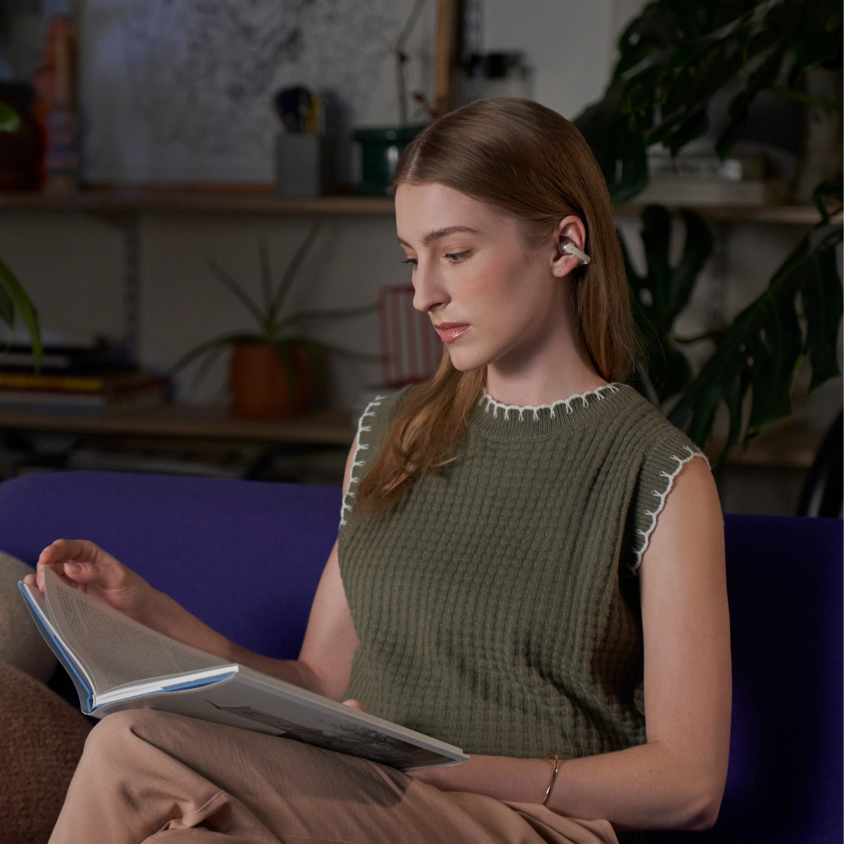 Kobieta w słuchawkach czyta książkę na fioletowej kanapie. Ma na sobie zielony top. Rośliny i półki w tle.
