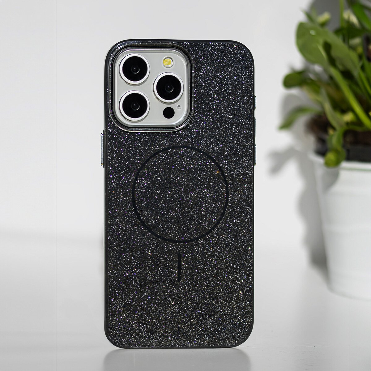 Schwarze Handyhülle mit Glitzer. Sie hat Kameraausschnitte und einen MagSafe-Kreis. Ein weißer Blumentopf ist im Hintergrund.