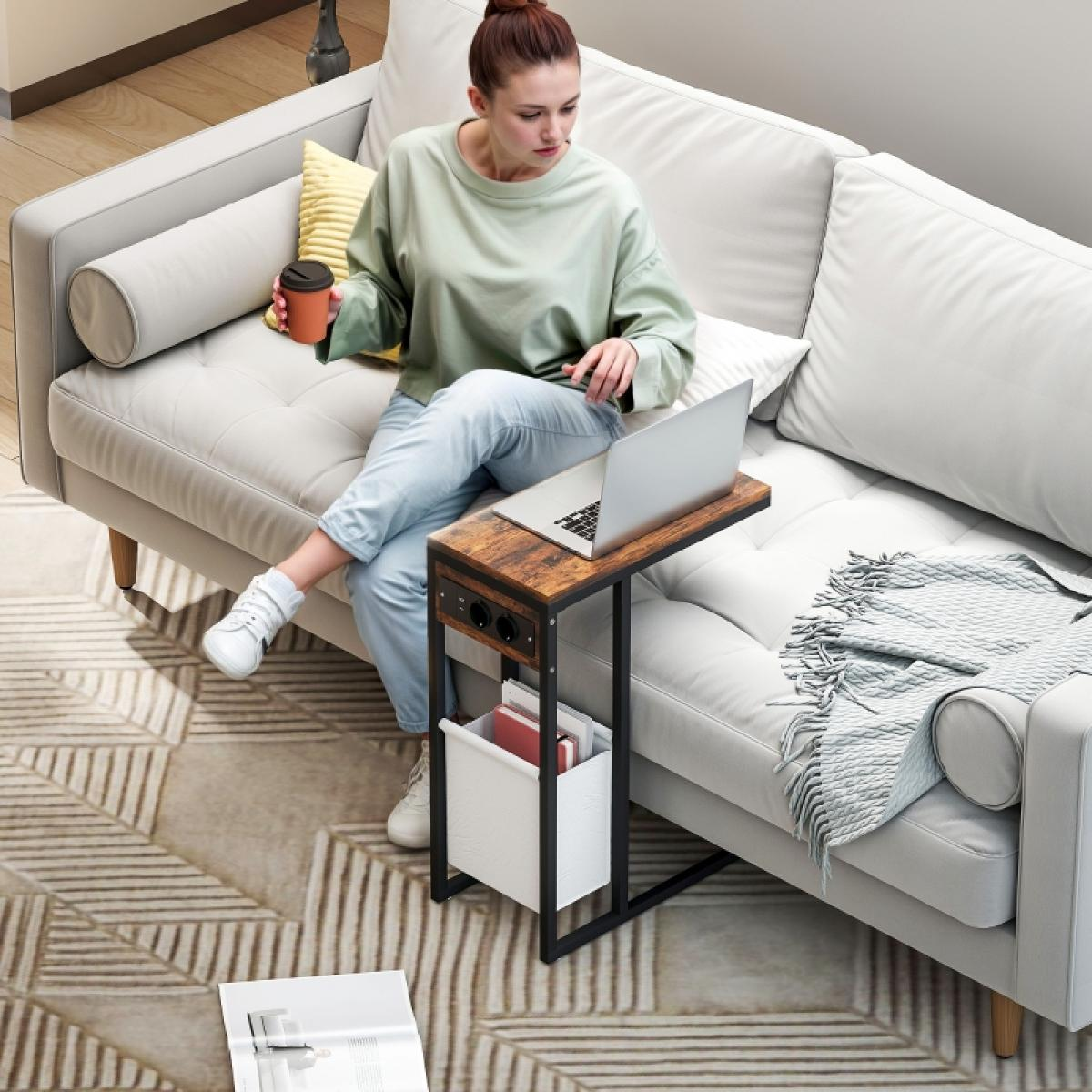 Eine Frau sitzt mit einem Laptop auf einem Sofa. Ein Beistelltisch hat eine Kaffeetasse und Ladeanschlüsse. Magazine und Decke sind sichtbar.