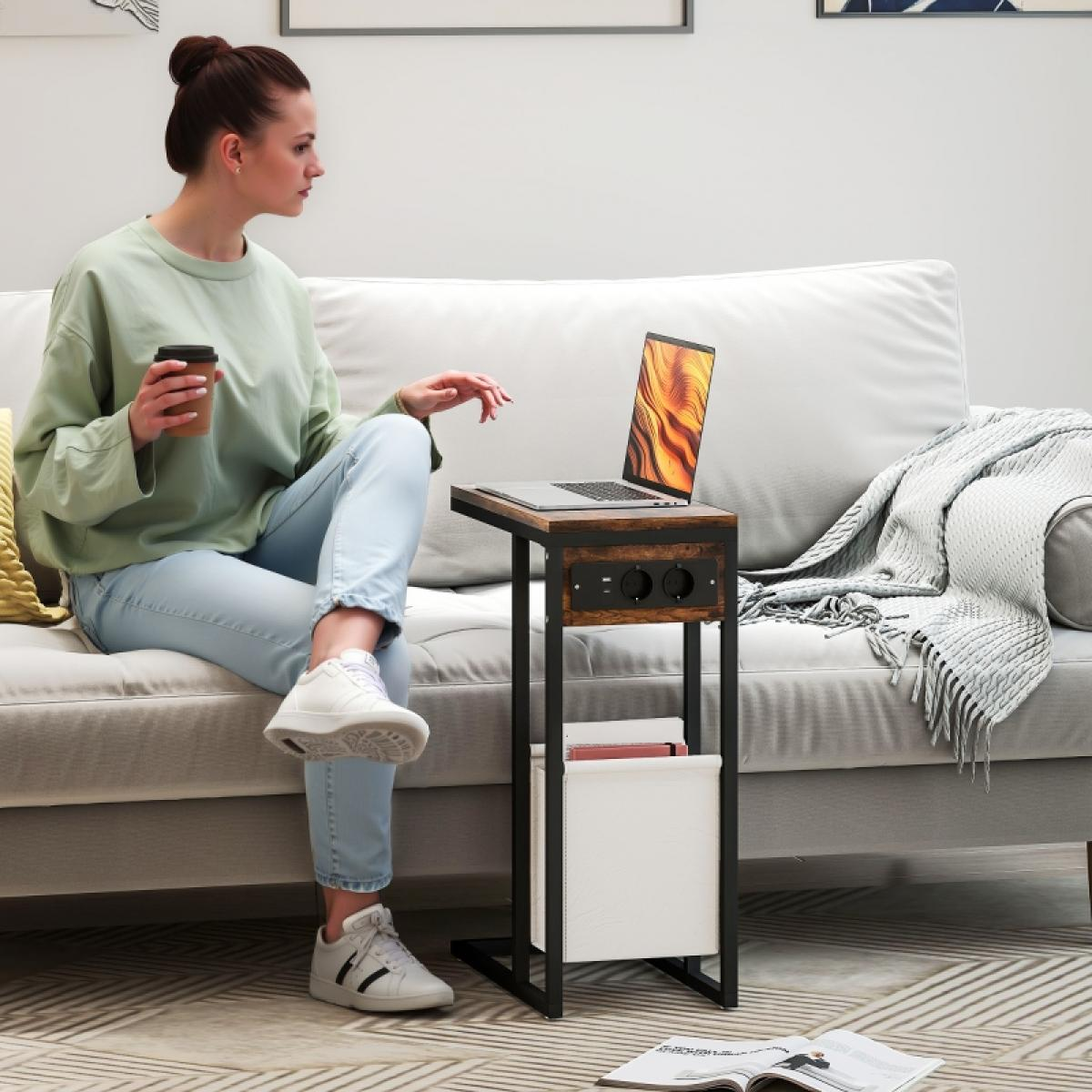 Frau auf Sofa mit Laptop auf Beistelltisch. Sie hält eine Tasse. Tisch hat Steckdosen und ein Magazinregal. Decken und ein Buch sind vorhanden.