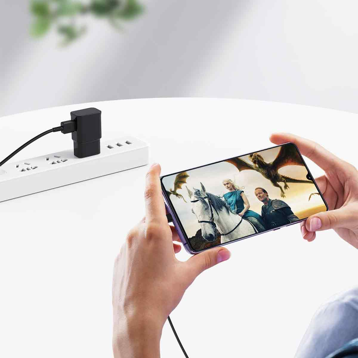 Hände halten ein Telefon, das eine Filmszene mit Figuren auf einem Pferd und Drachen zeigt. Ein Netzteil ist in eine Steckdosenleiste eingesteckt.
