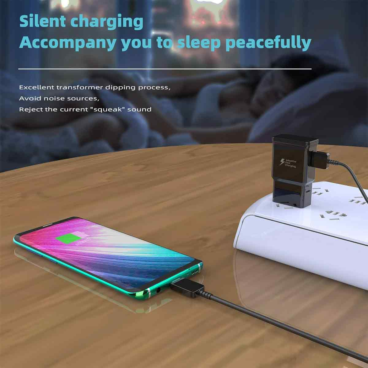 Ein Telefon, das auf einem Holztisch geladen wird. Ein schwarzes Ladegerät und eine Steckdosenleiste sind sichtbar. Der Hintergrund zeigt eine schlafende Person.