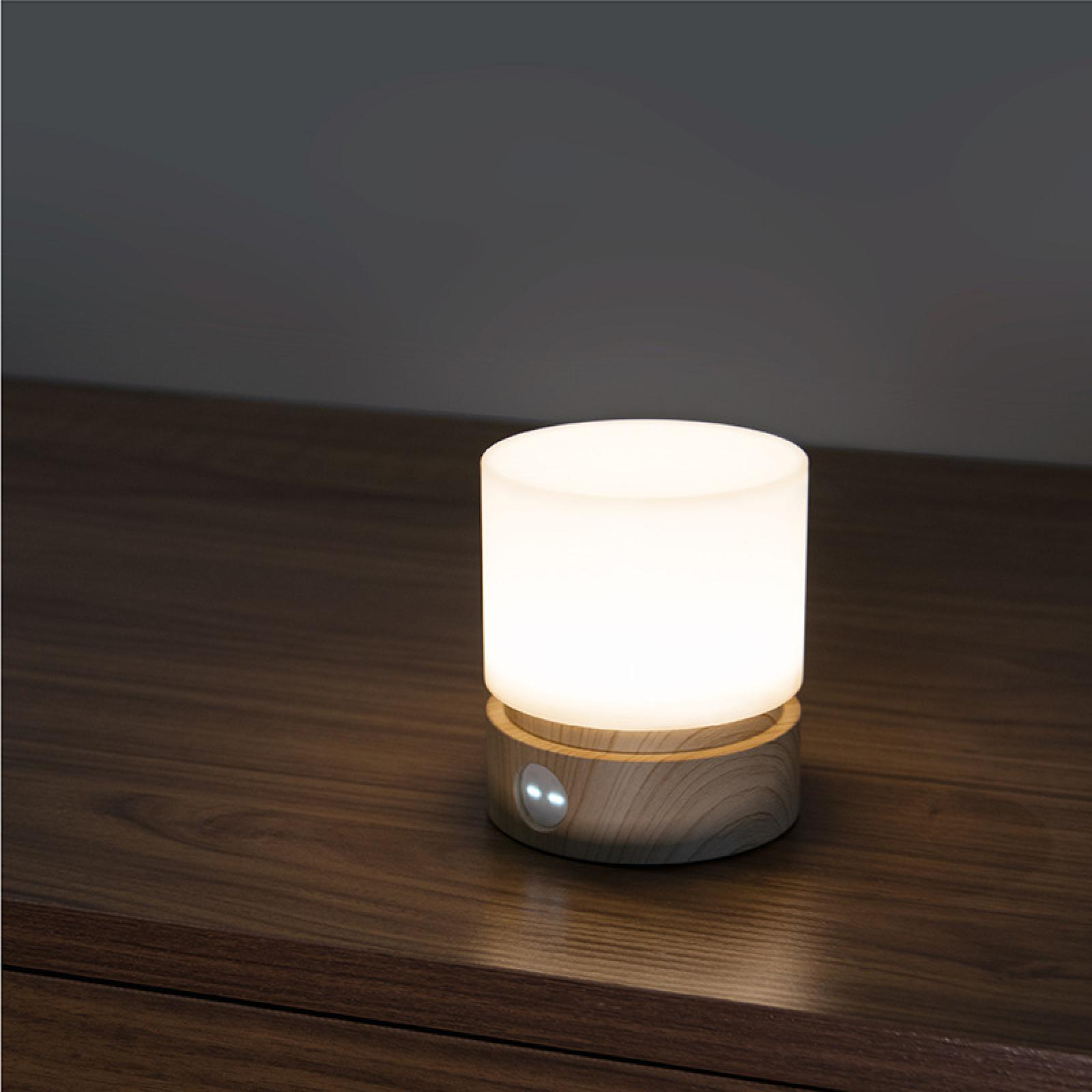 Eine beleuchtete, zylindrische Lampe steht auf einer dunklen Holzoberfläche. Die Lampe hat einen Holzfuß und eine weiße, leuchtende Oberseite.