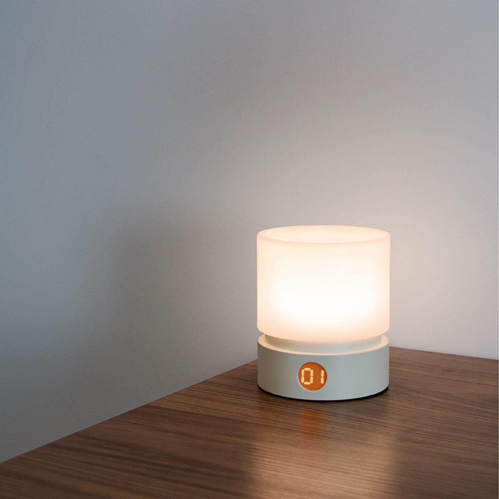 Eine leuchtende Nachttischlampe auf einer Holzoberfläche. Die Lampe hat einen weißen Sockel, eine leuchtende Oberseite und zeigt die Zahl '01'.