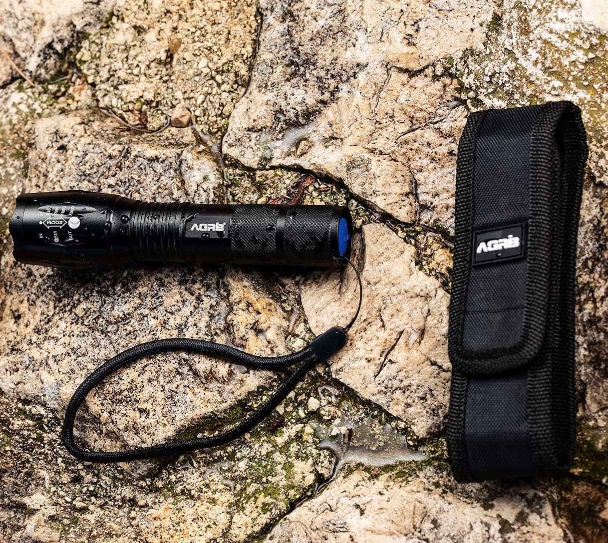 Eine schwarze Taschenlampe, ihre Halterung und ein Lanyard liegen auf einer strukturierten Felsfläche. Die Taschenlampe hat blaue Akzente und den Markennamen AGRIS.