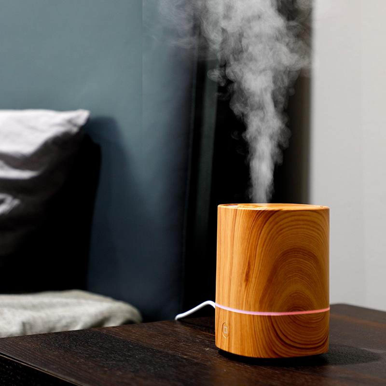 Ein hölzerner Aromadiffusor gibt Dampf ab. Er steht auf einem dunklen Tisch, mit einem verschwommenen Hintergrund aus Wand und Kissen.