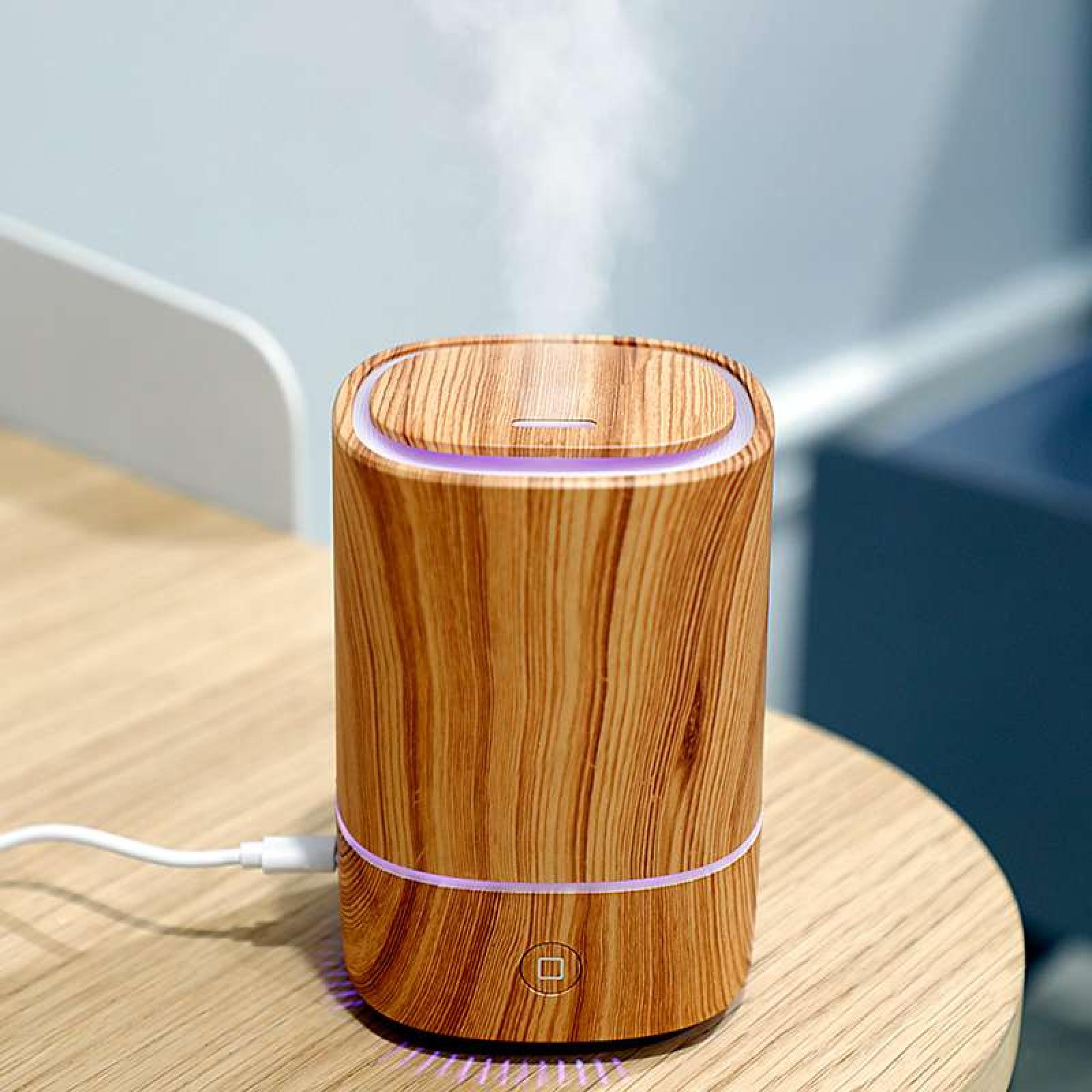 Ein Diffusor in Holzoptik mit lila Licht. Er steht auf einem Holztisch. Dampf steigt auf. Ein weißes Kabel ist eingesteckt.